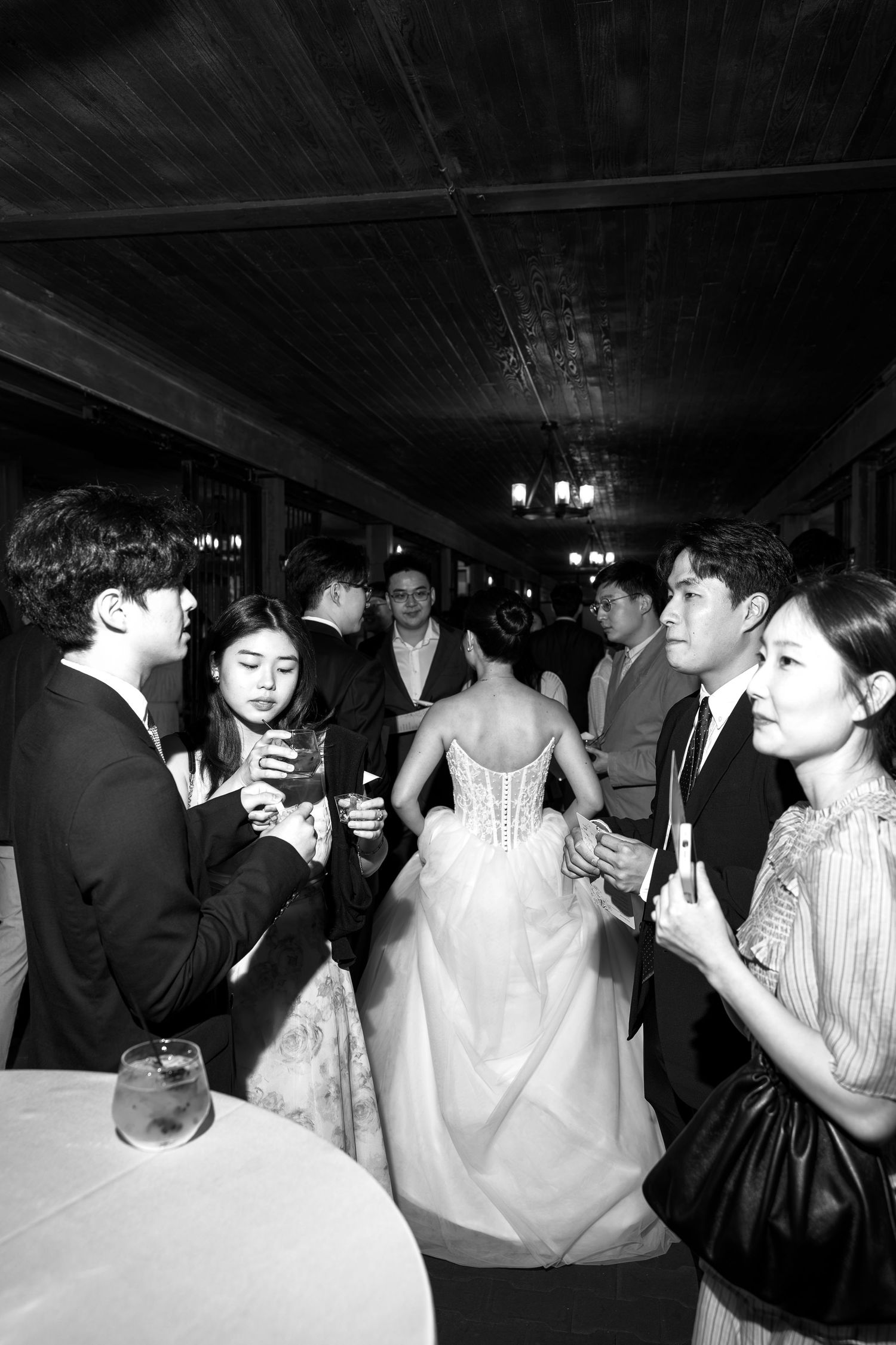 Black and white photo of wedding guests mingling at a reception, bride in white gown visible among elegantly dressed attendees.