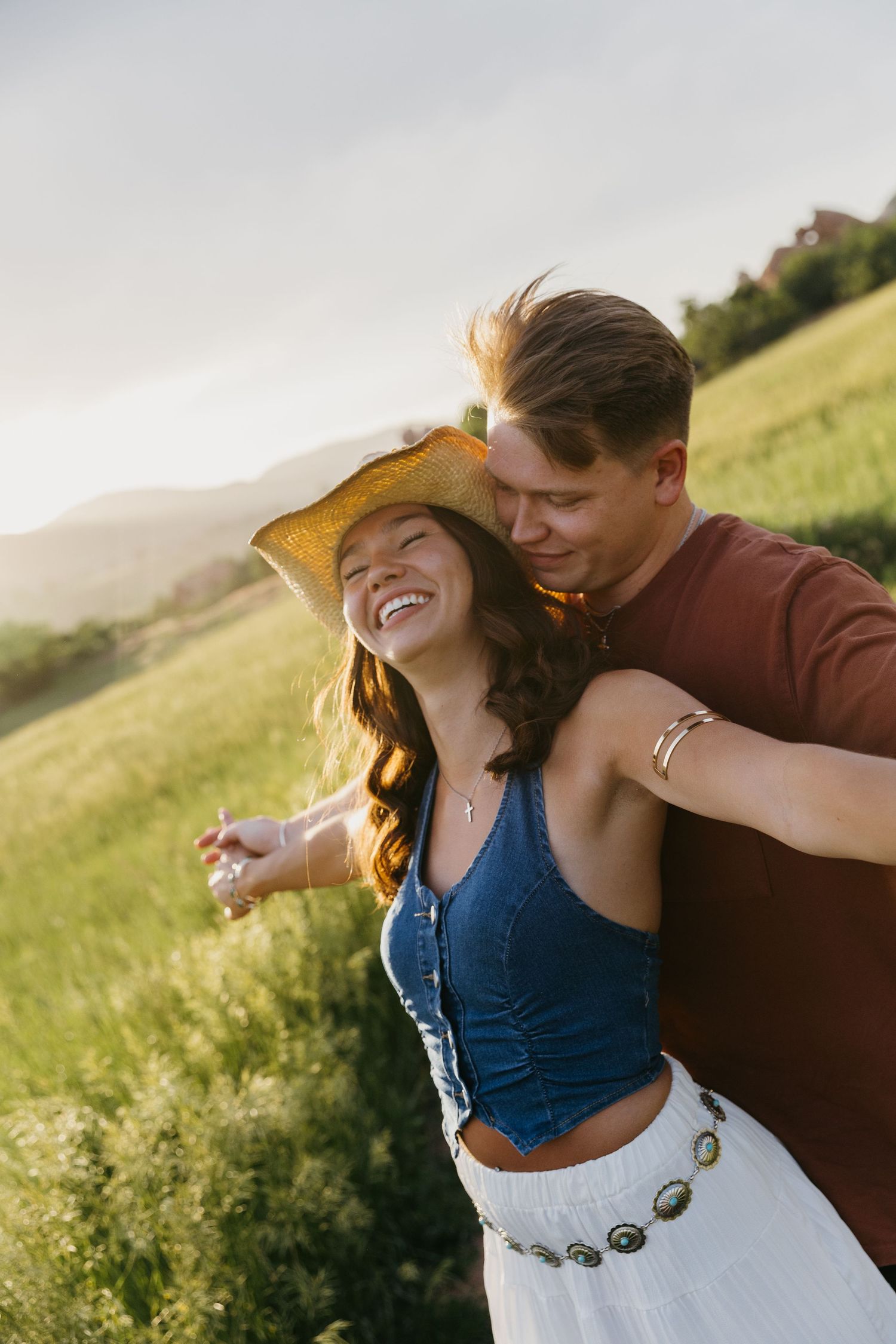 A laughing woman in a cowboy hat is embraced from behind by her partner in a sunlit green field.