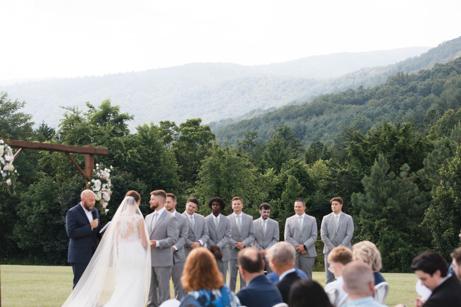 Outdoor wedding ceremony with mountain views, groomsmen in gray suits, and bride and groom at rustic wooden arch altar.
