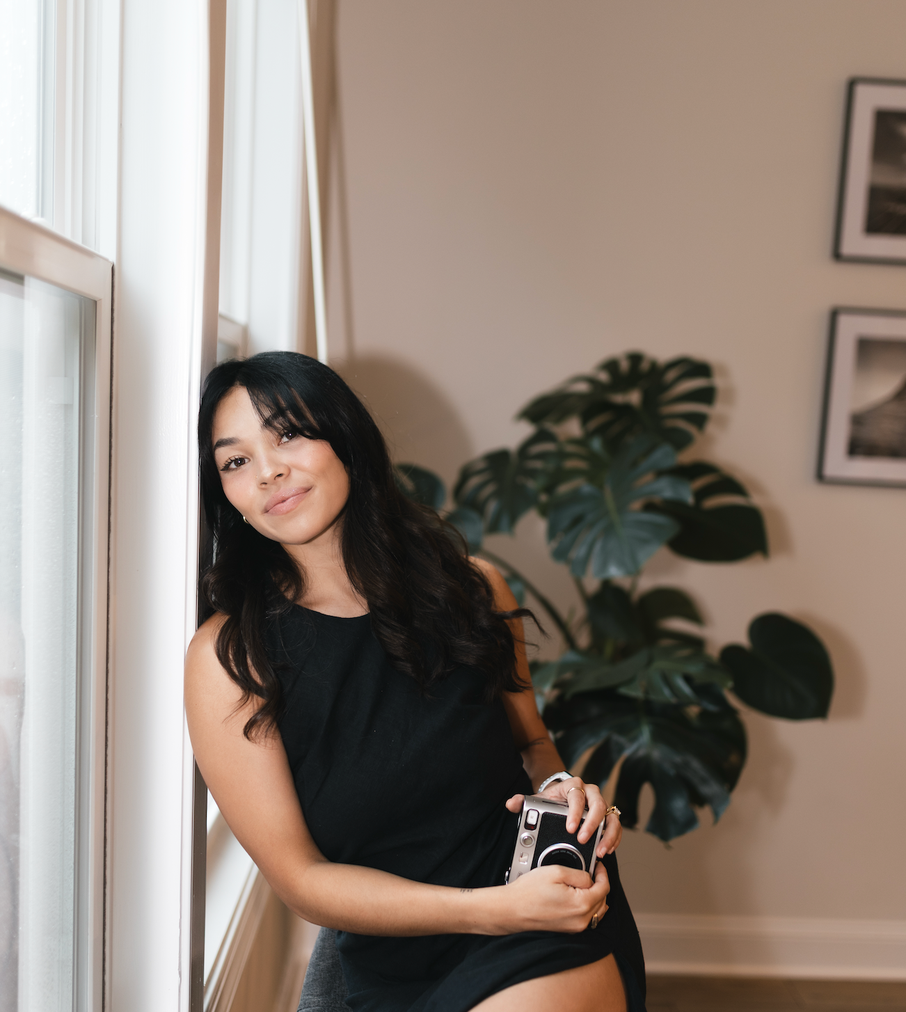 Professional wedding photographer in black dress posing with camera near window and monstera plant.