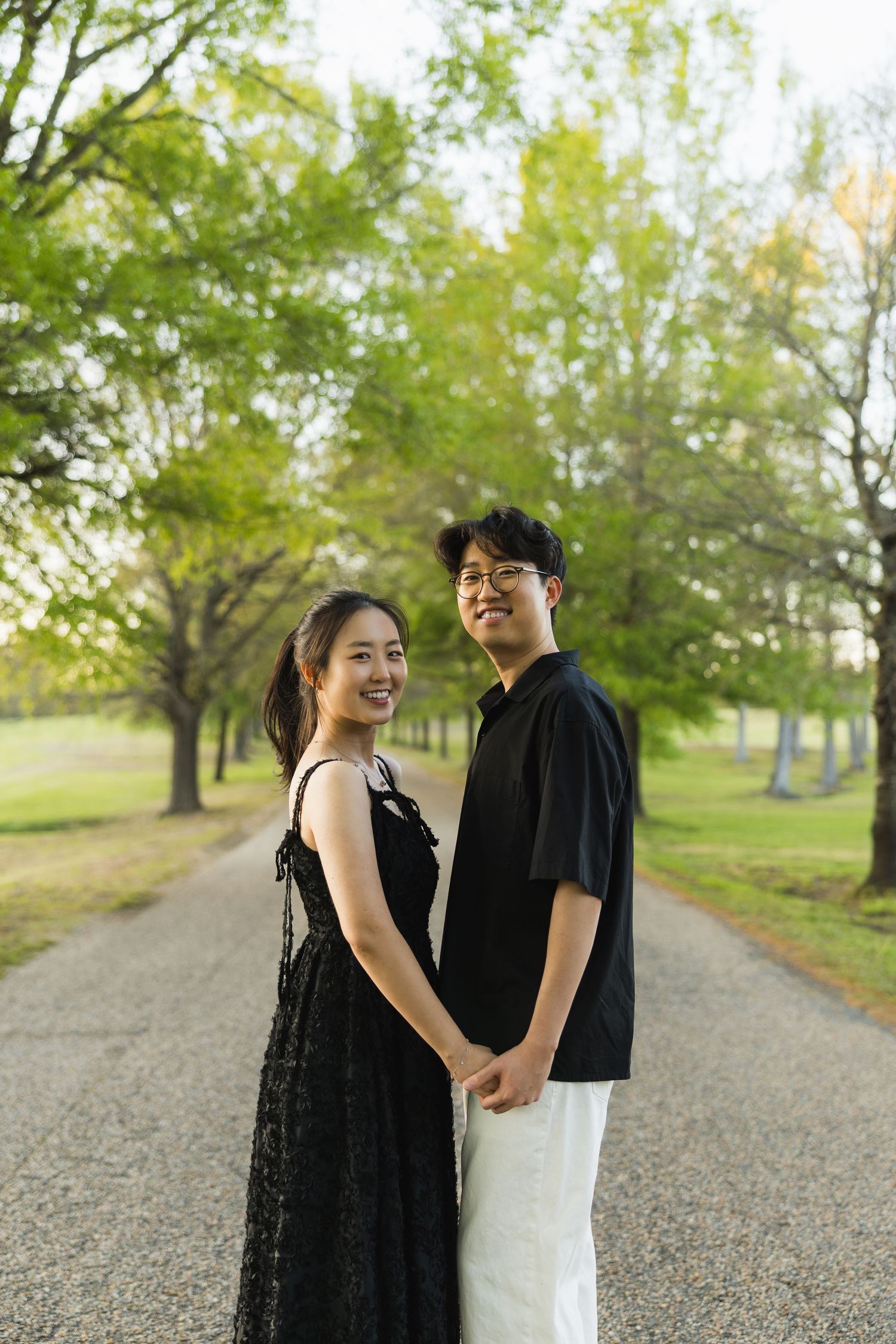 A smiling couple holds hands on a tree-lined park path, both dressed in black, posing for an engagement photo.