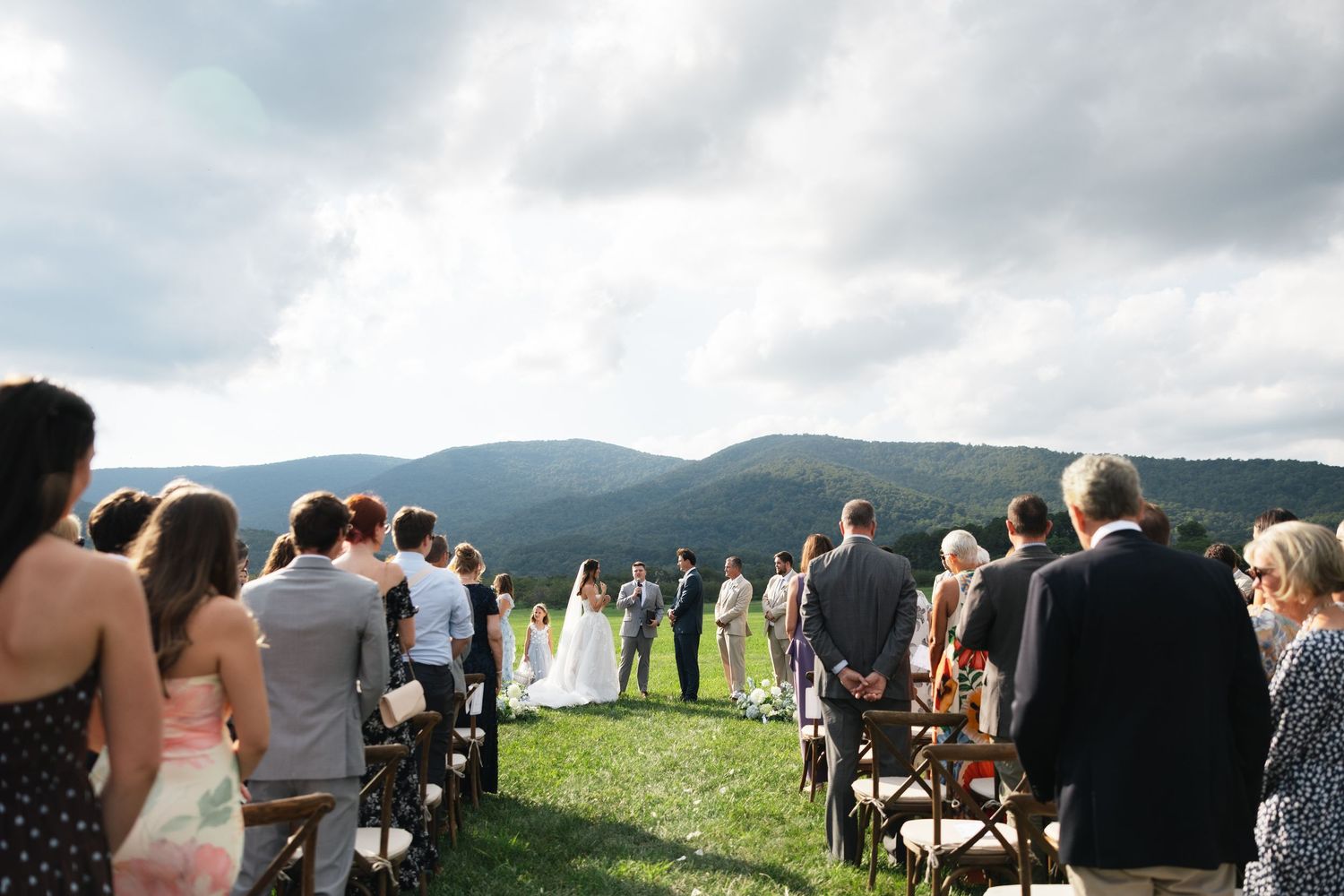 Outdoor wedding ceremony on a hilltop with mountain views, guests seated in wooden chairs under a cloudy sky.