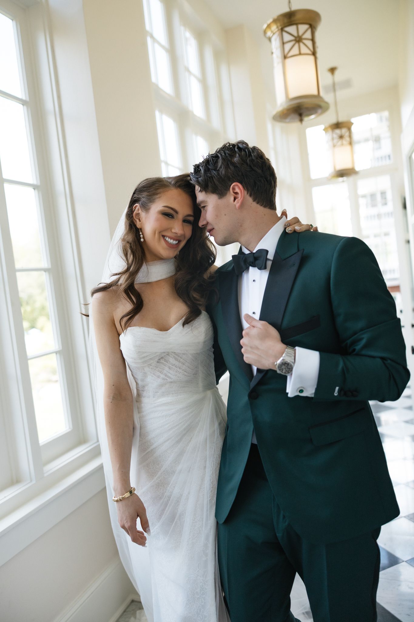 A bride in a white gown smiles as her groom in a dark green tuxedo whispers in her ear in an elegant venue.