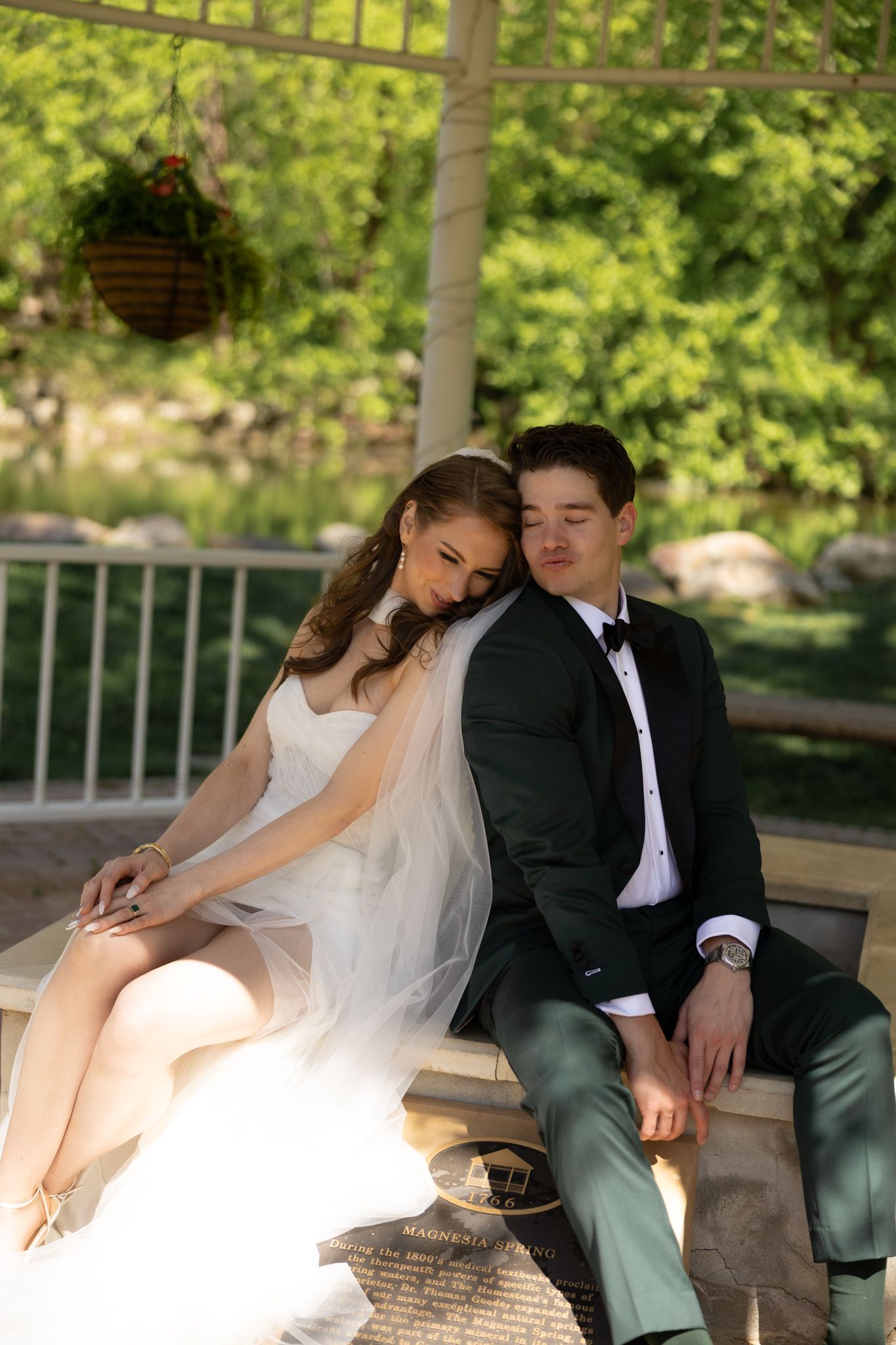 Bride and groom in wedding attire sitting closely together on a bench in a garden gazebo.