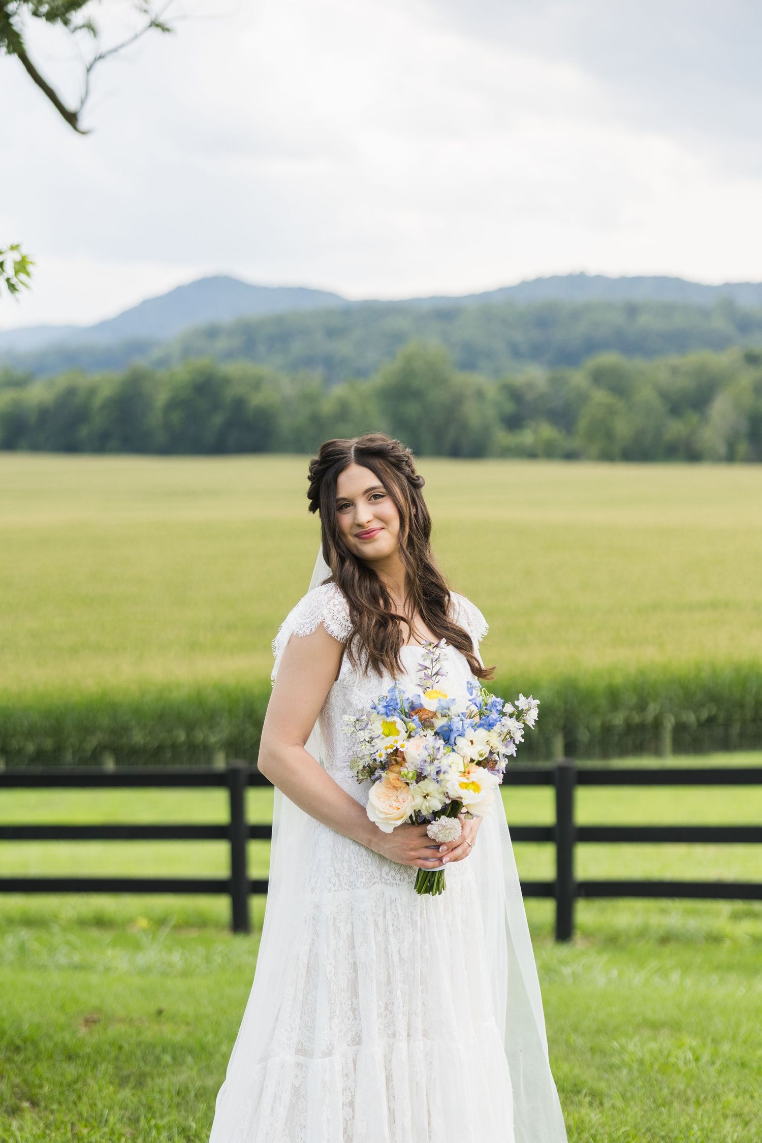Bride in white lace gown holding blue and white bouquet standing in front of fence with mountain backdrop.
