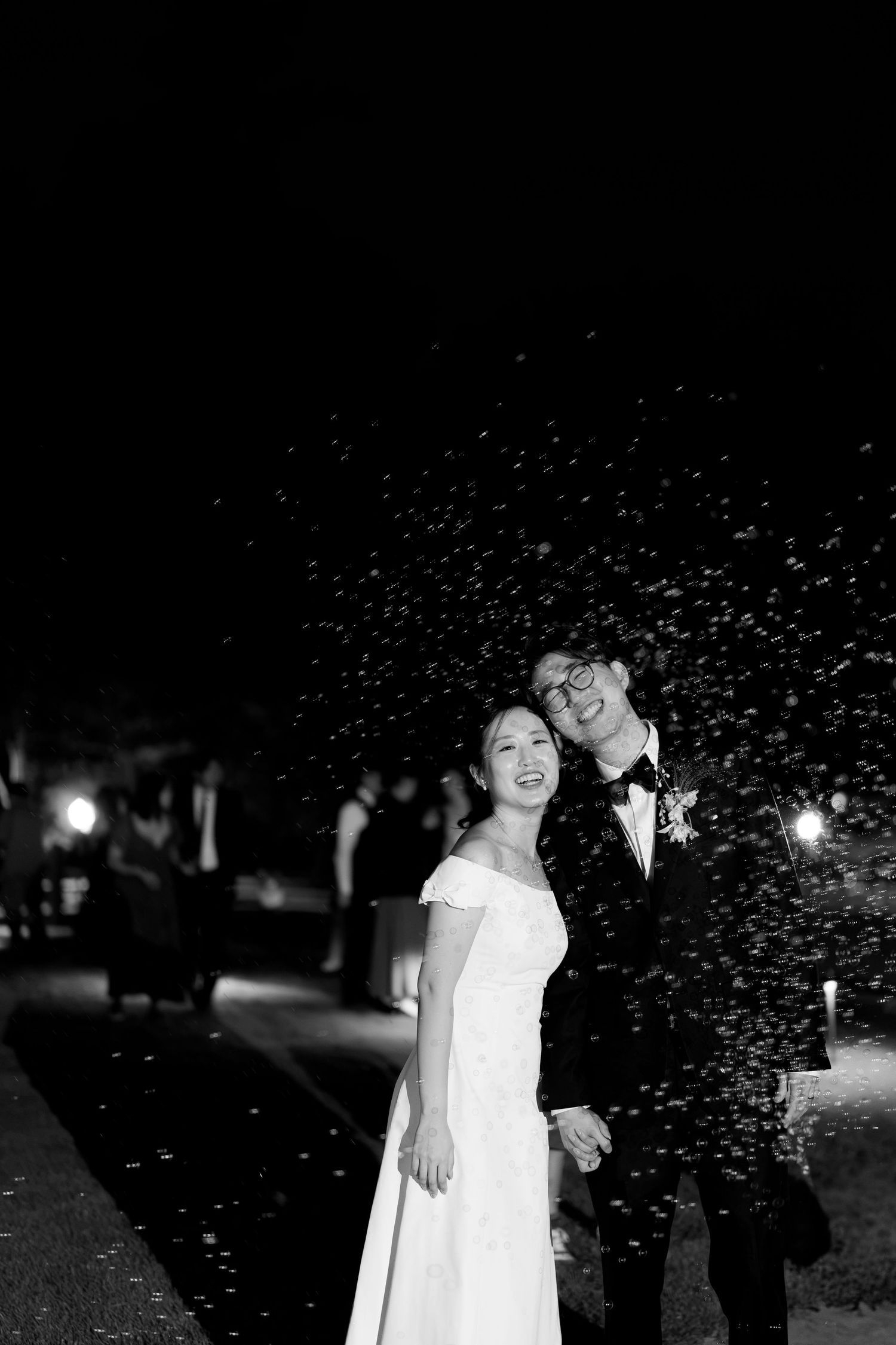Black and white photo of a smiling couple in wedding attire celebrating outdoors at night with confetti falling around them.