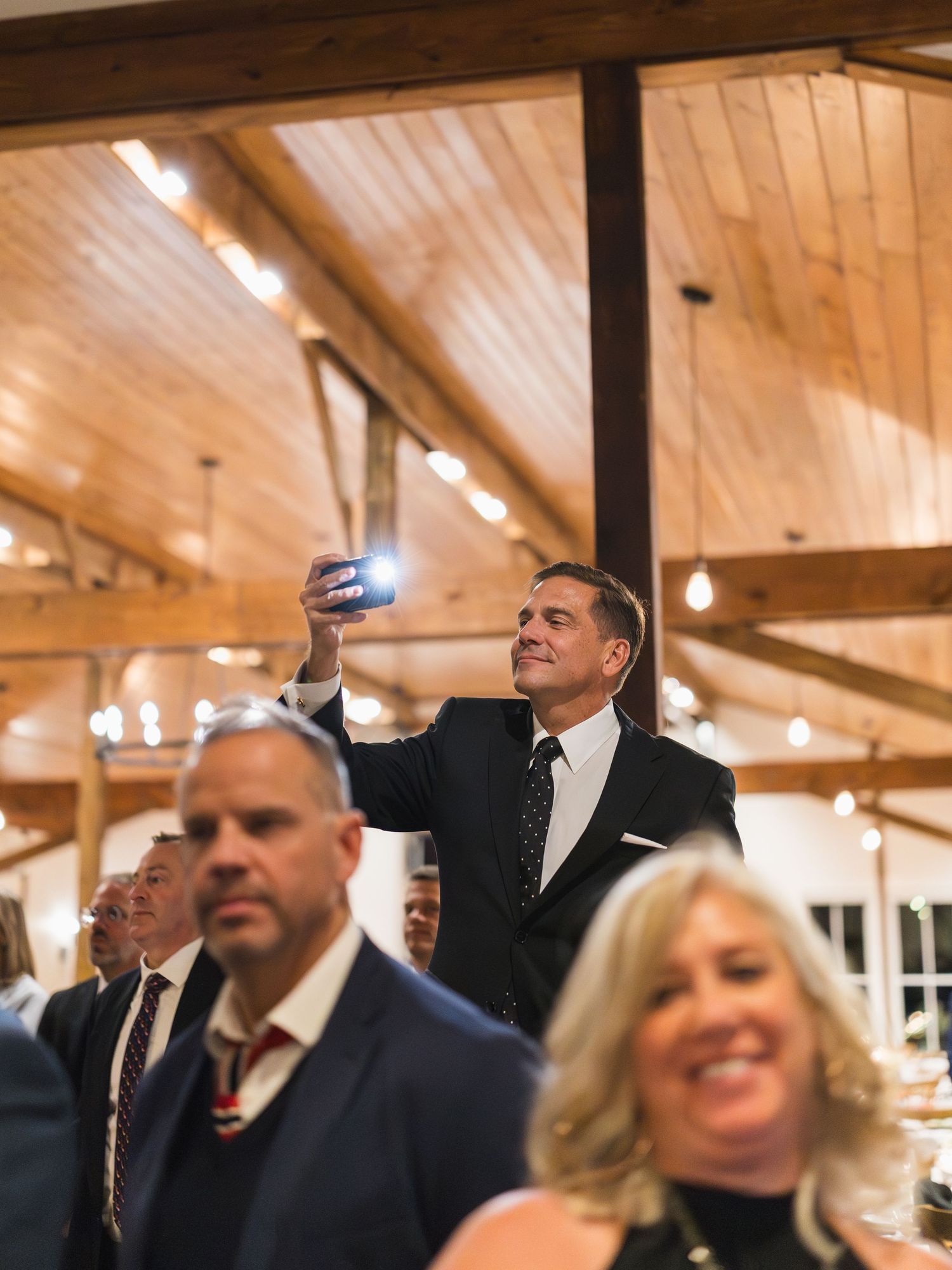 Wedding guest holds up smartphone to capture a photo inside a rustic barn reception venue with string lights.