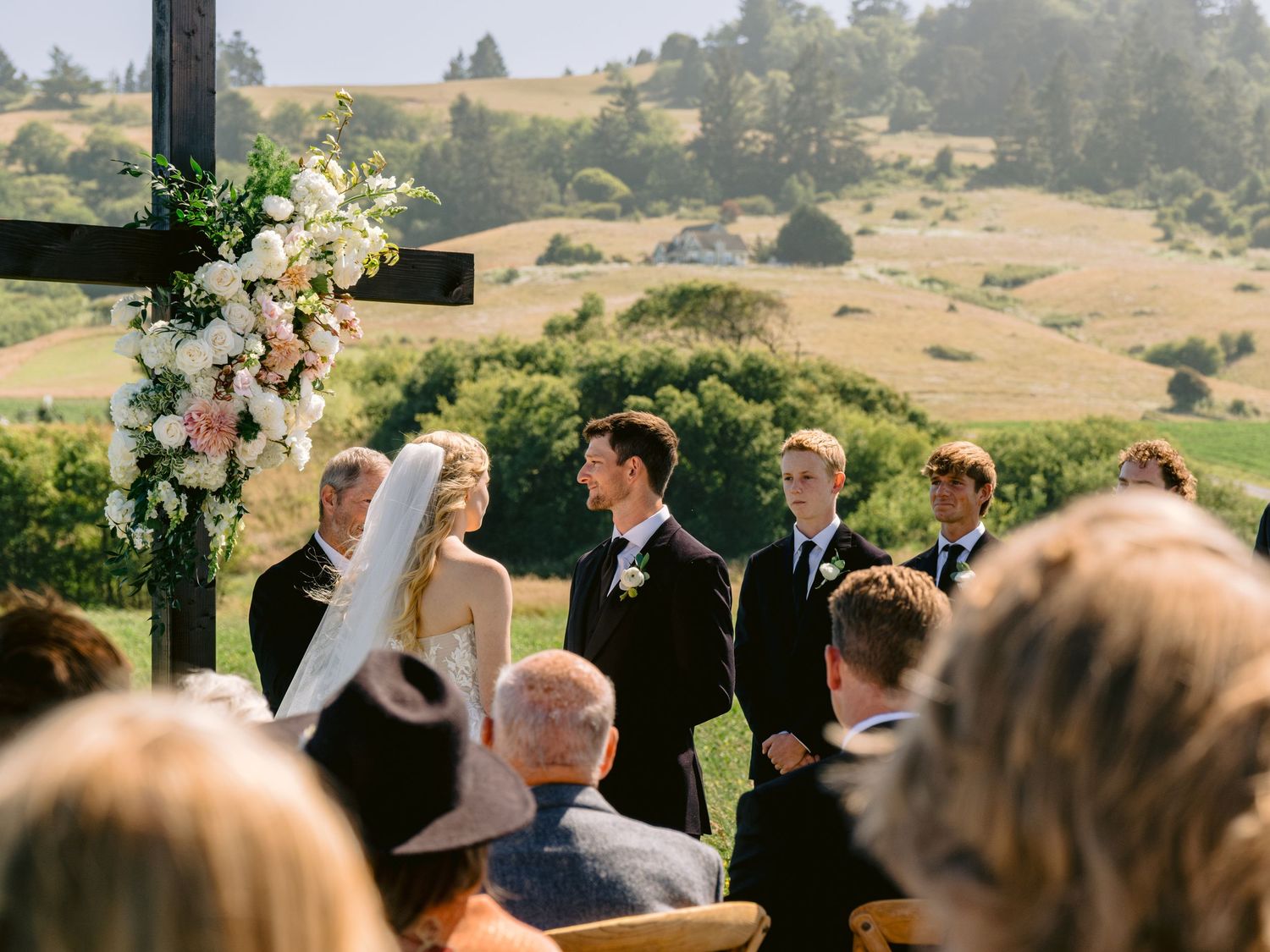 Outdoor wedding ceremony with guests gathered under a covered pavilion overlooking rolling hills.