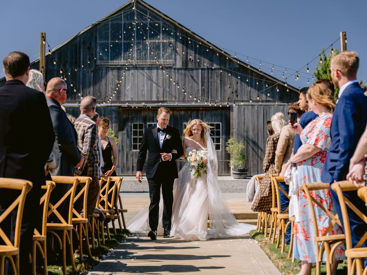 Wedding ceremony aisle with wooden chairs lined up leading to a modern barn structure.