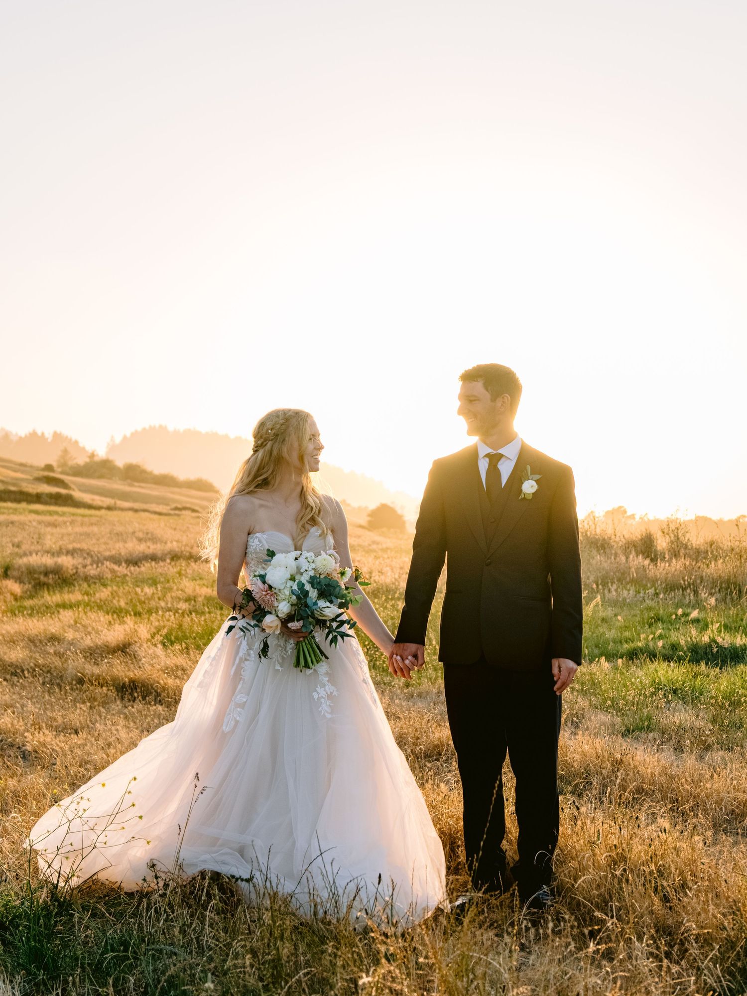 Wedding couple walks through a sun-drenched field during golden hour photography session.