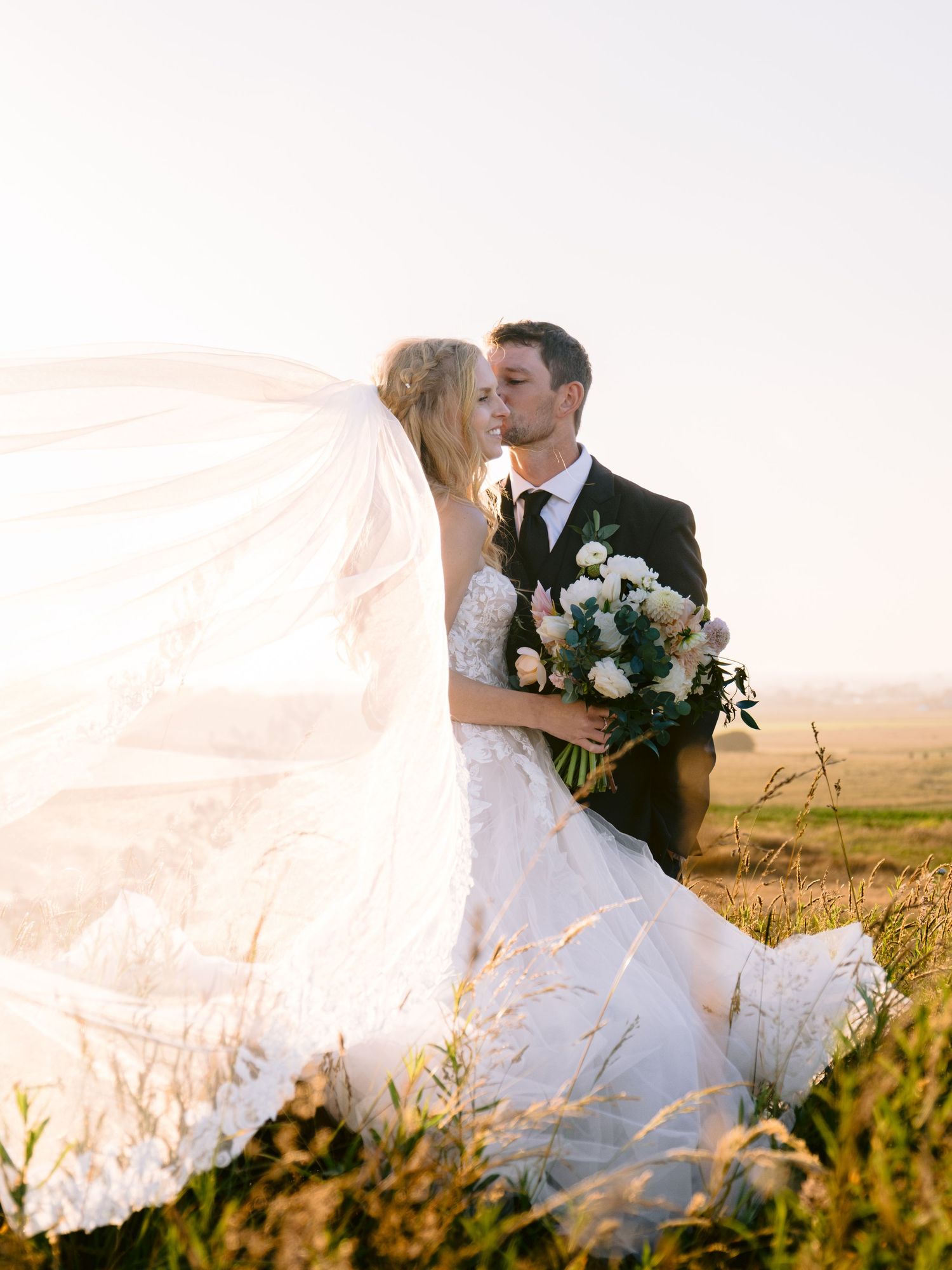 A romantic wedding couple embraces in a golden wheat field at sunset with a flowing veil in the breeze.