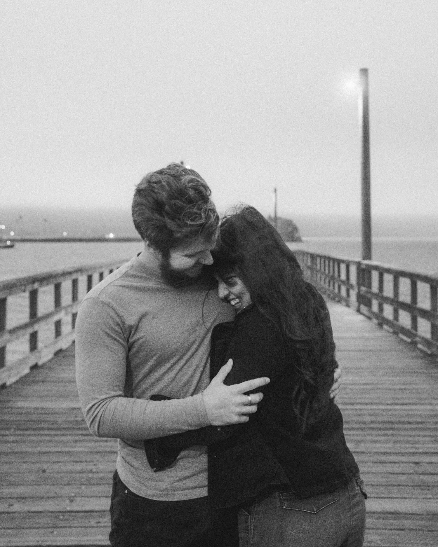 Couple embracing on wooden pier with foggy ocean backdrop in black and white.