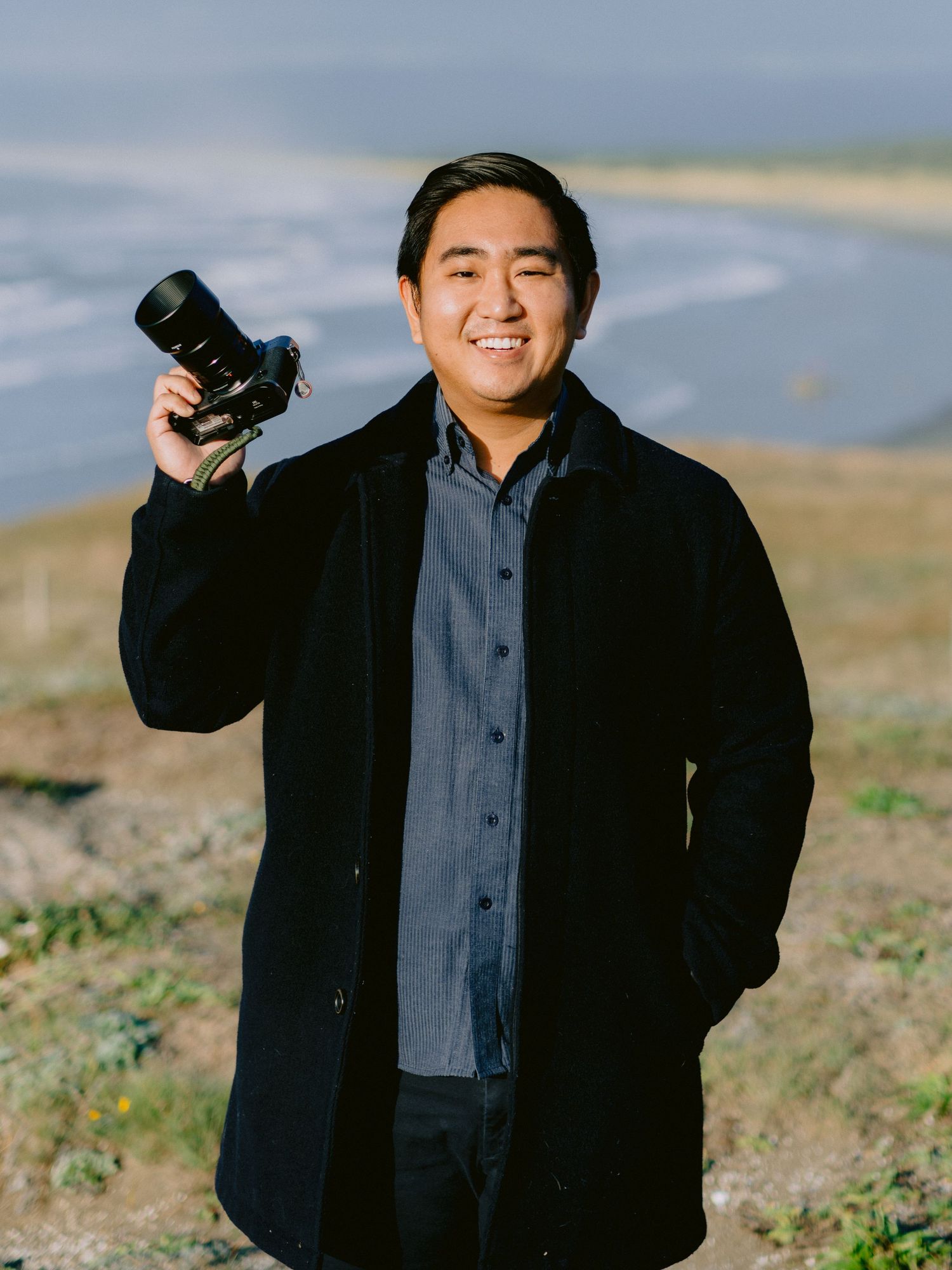 A photographer in a black coat holds up their camera while posing against a scenic outdoor backdrop.