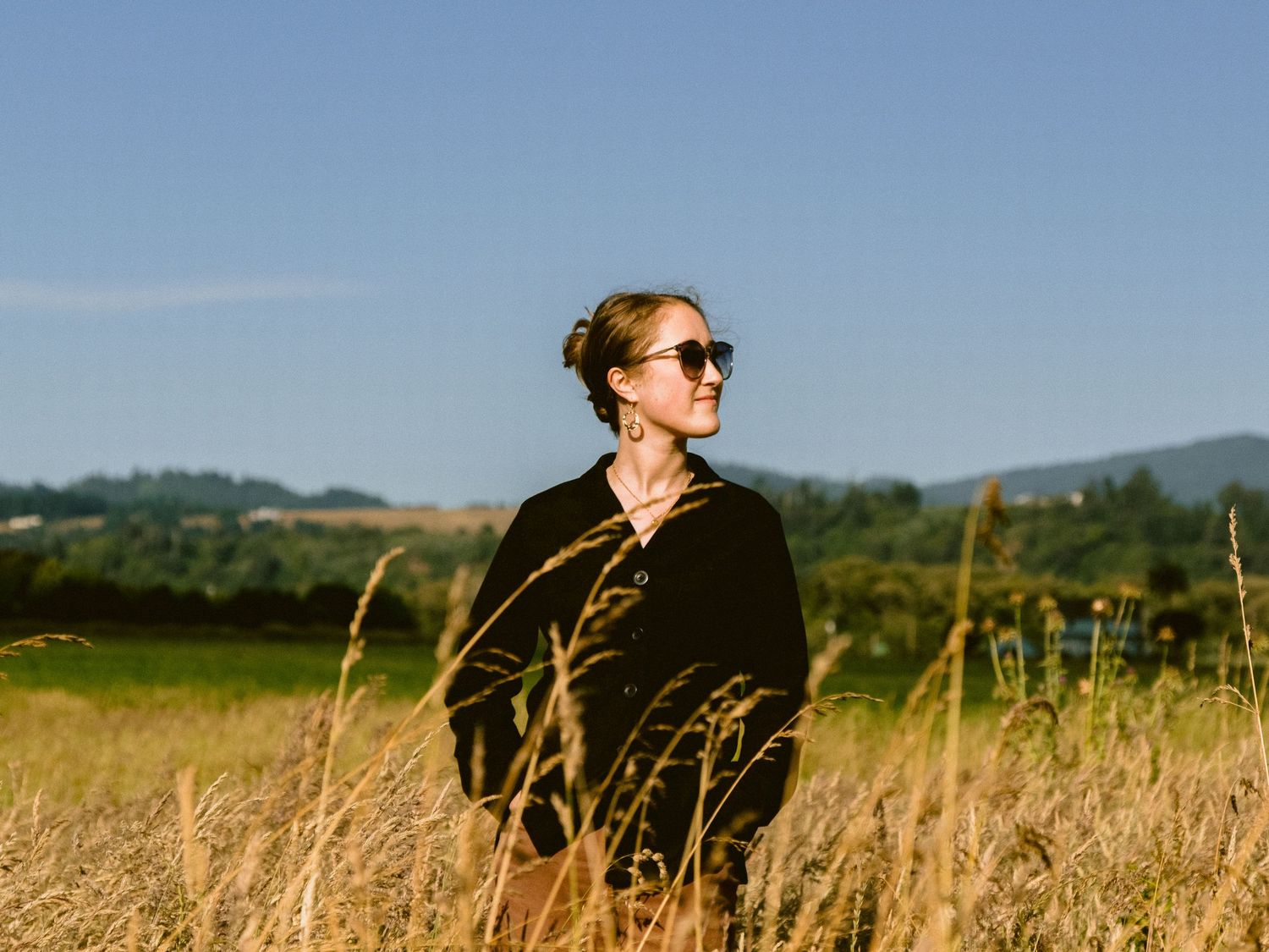 Figure in black clothing standing in tall golden grass with hills in background.