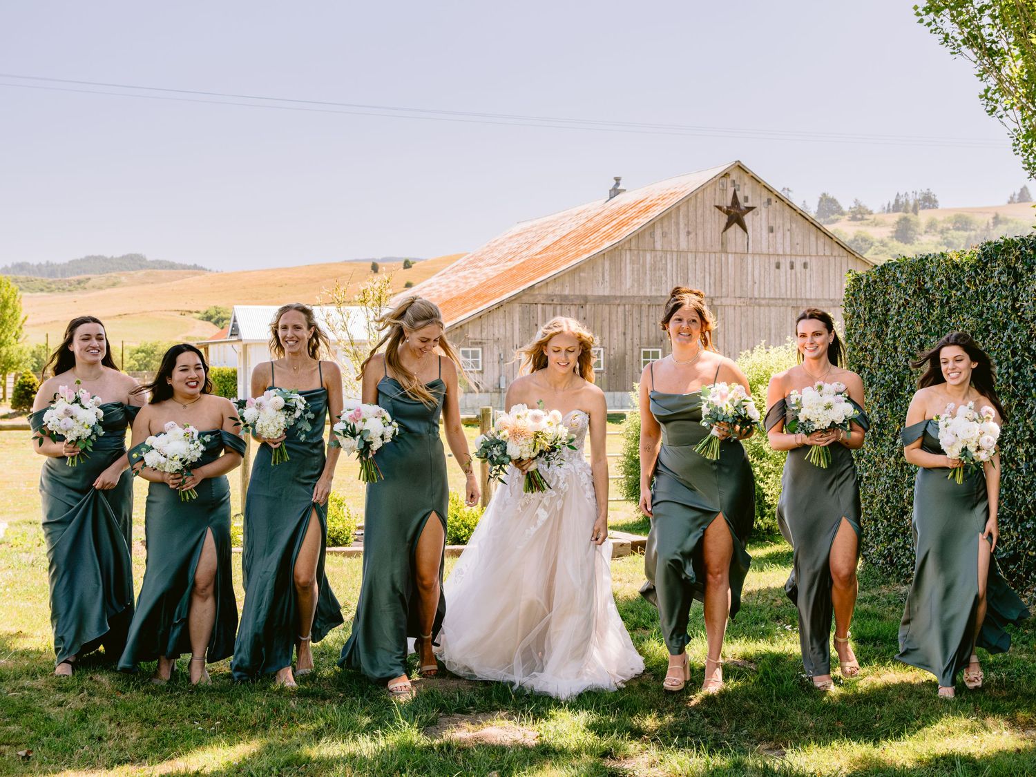 Wedding party in green dresses posing playfully on grass with rustic barn in background.