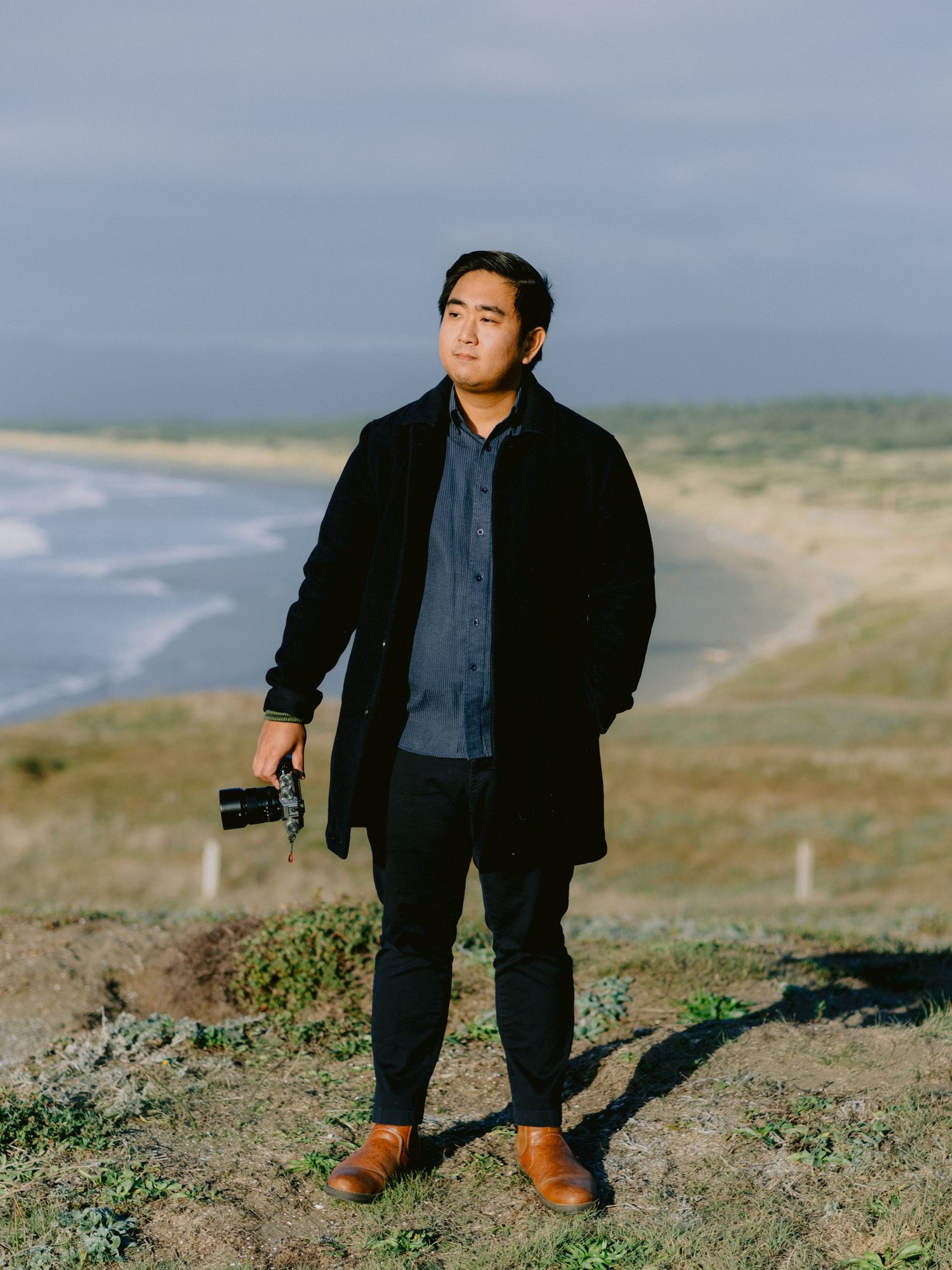 A person in a black suit and brown shoes stands on a coastal hillside overlooking the ocean.