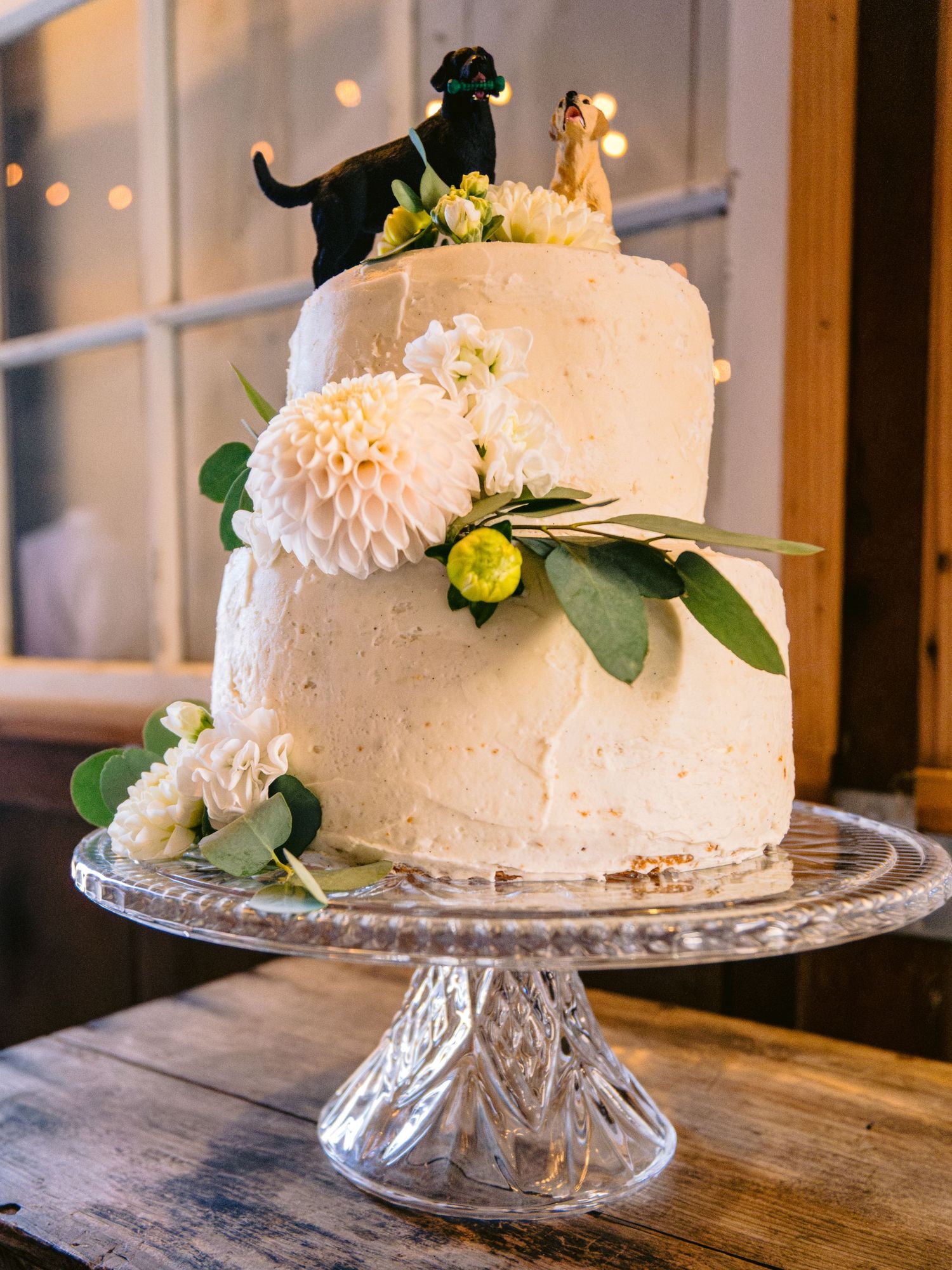 Elegant white wedding cake with fresh flowers and greenery on crystal cake stand.