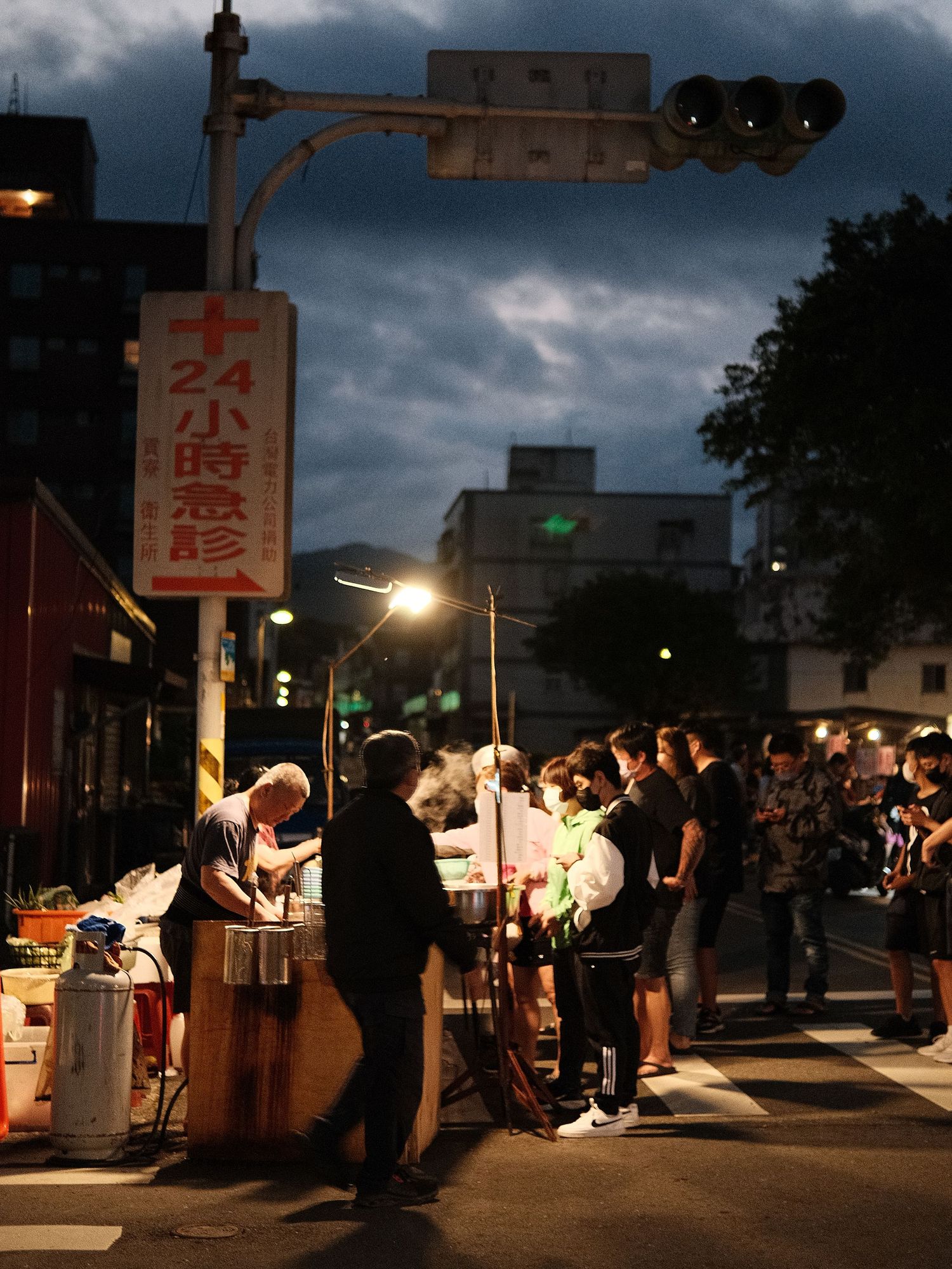 People gathering at an outdoor night market with food stalls under dark cloudy skies.