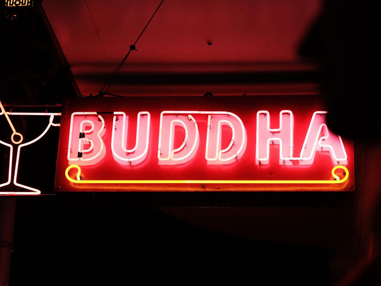 Red neon Buddha sign with cocktail glass icon glowing against dark background.