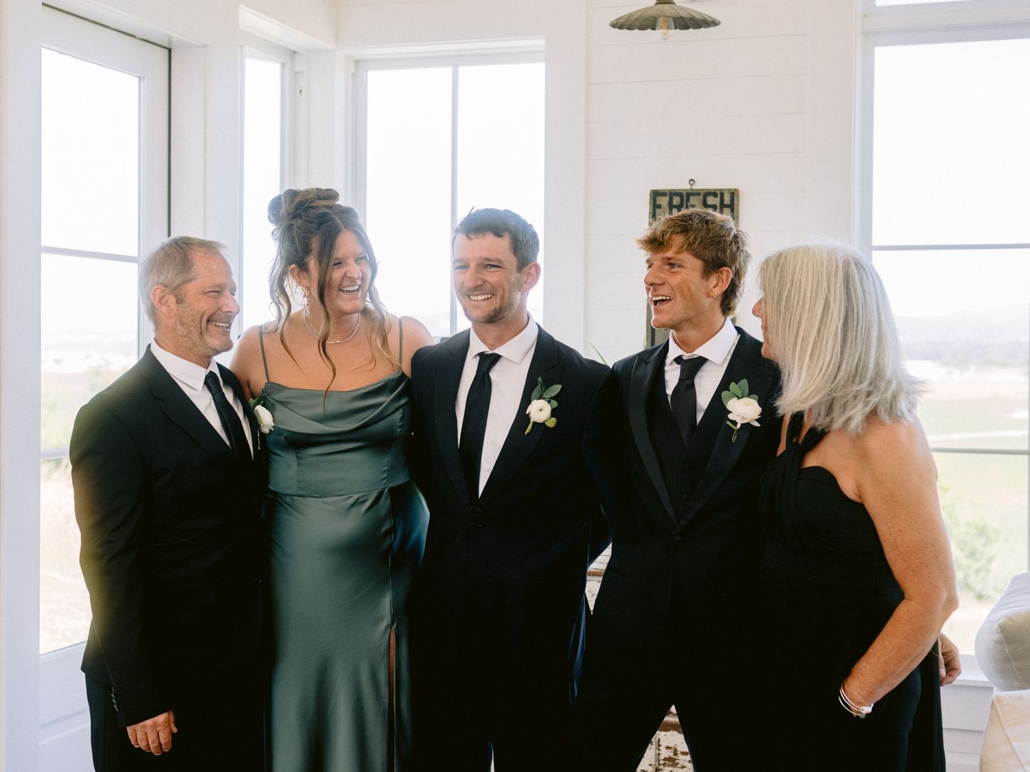 Wedding party in formal black attire poses together indoors by large windows.