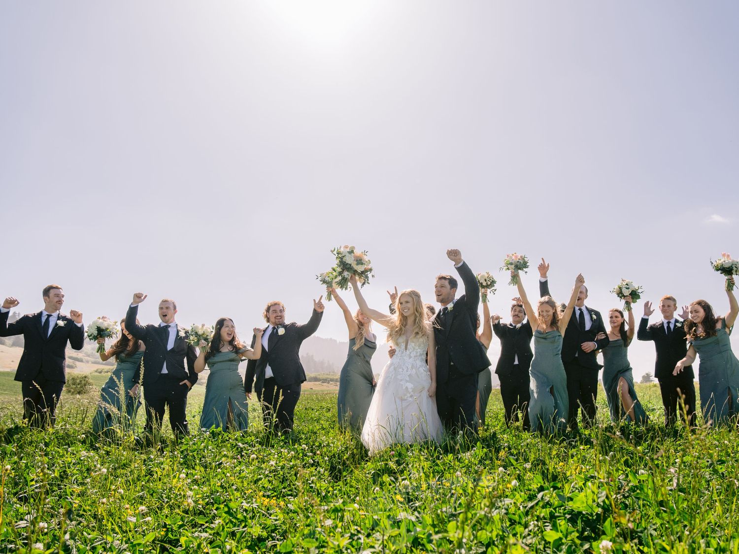 Wedding party and guests celebrating with raised arms against a scenic countryside backdrop.