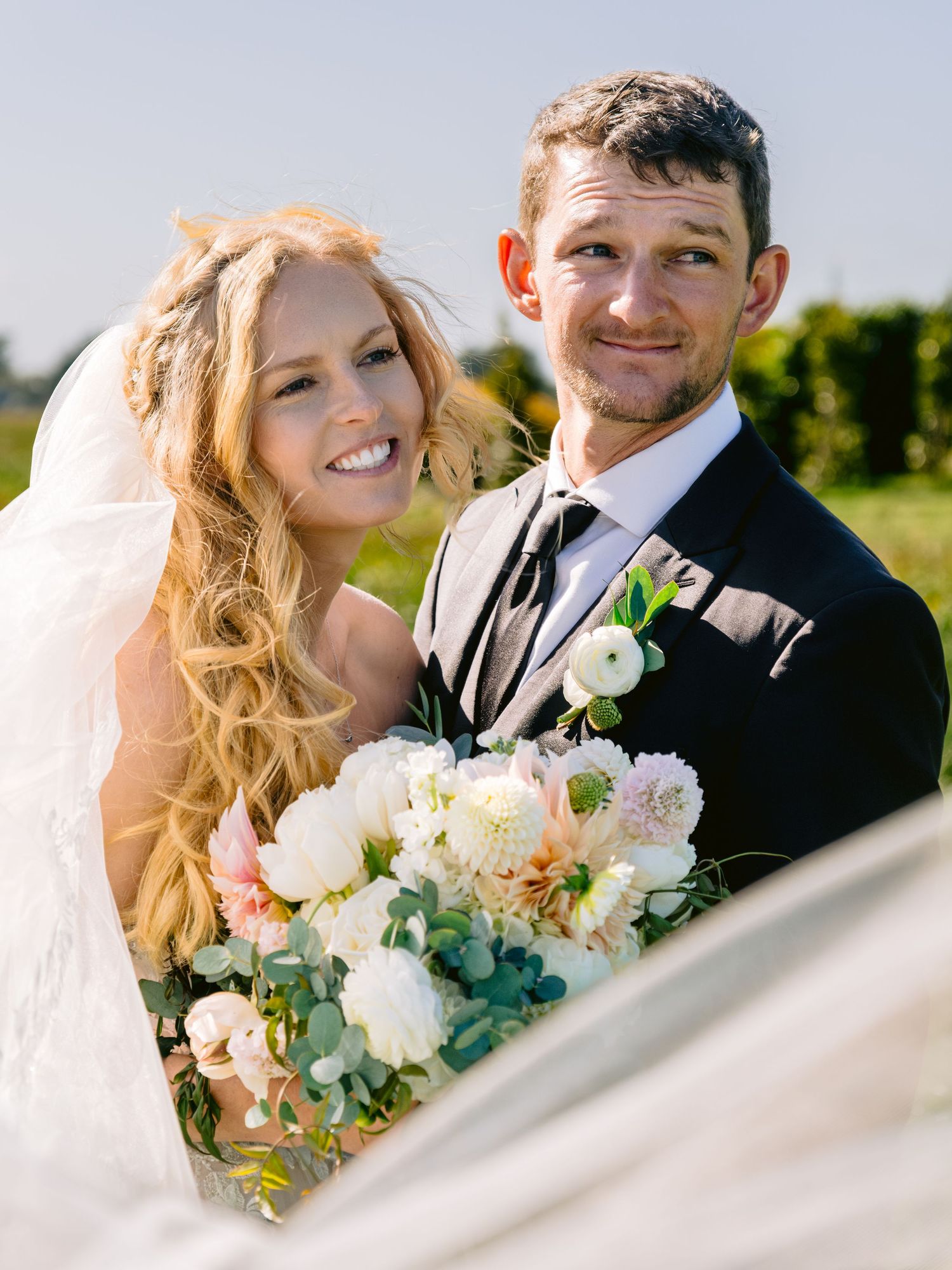 Wedding couple poses with a beautiful bouquet of white and peach flowers against a bright sky.