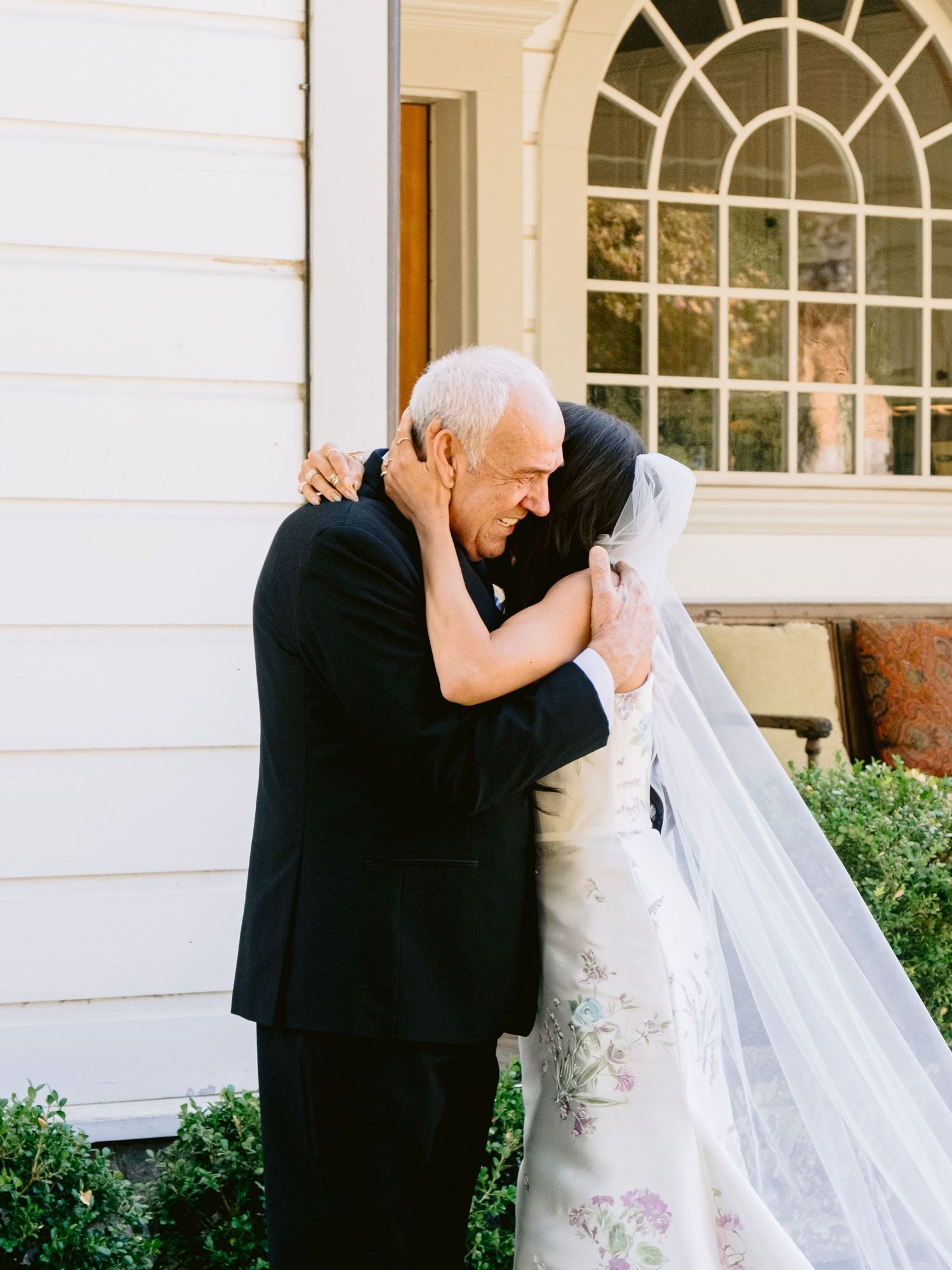 A tender moment between a bride and loved one outside a white building with arched windows.