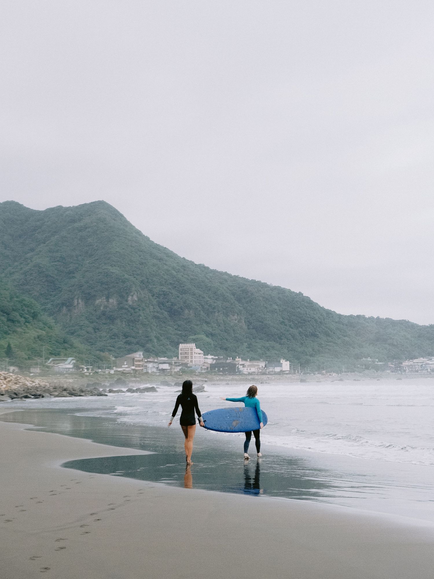 Surfers walking along a misty beach with mountains in the background.