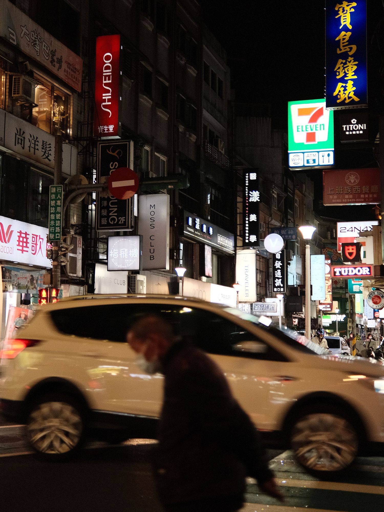 Blurred motion of cars driving past neon signs and storefronts at night in an Asian city.