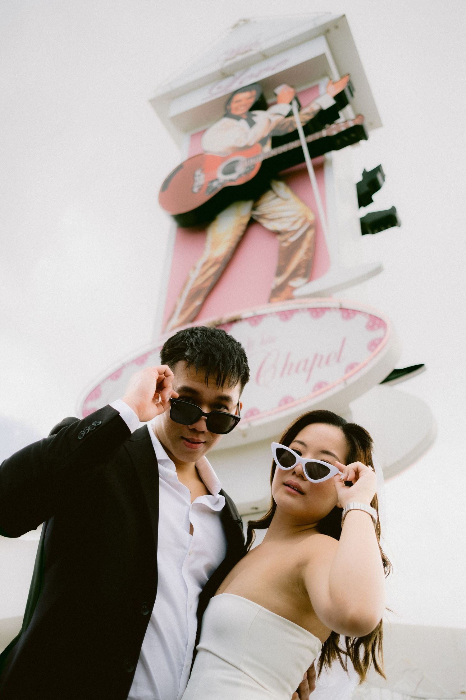 Couple in formal attire wearing retro sunglasses share a romantic moment outside a chapel.