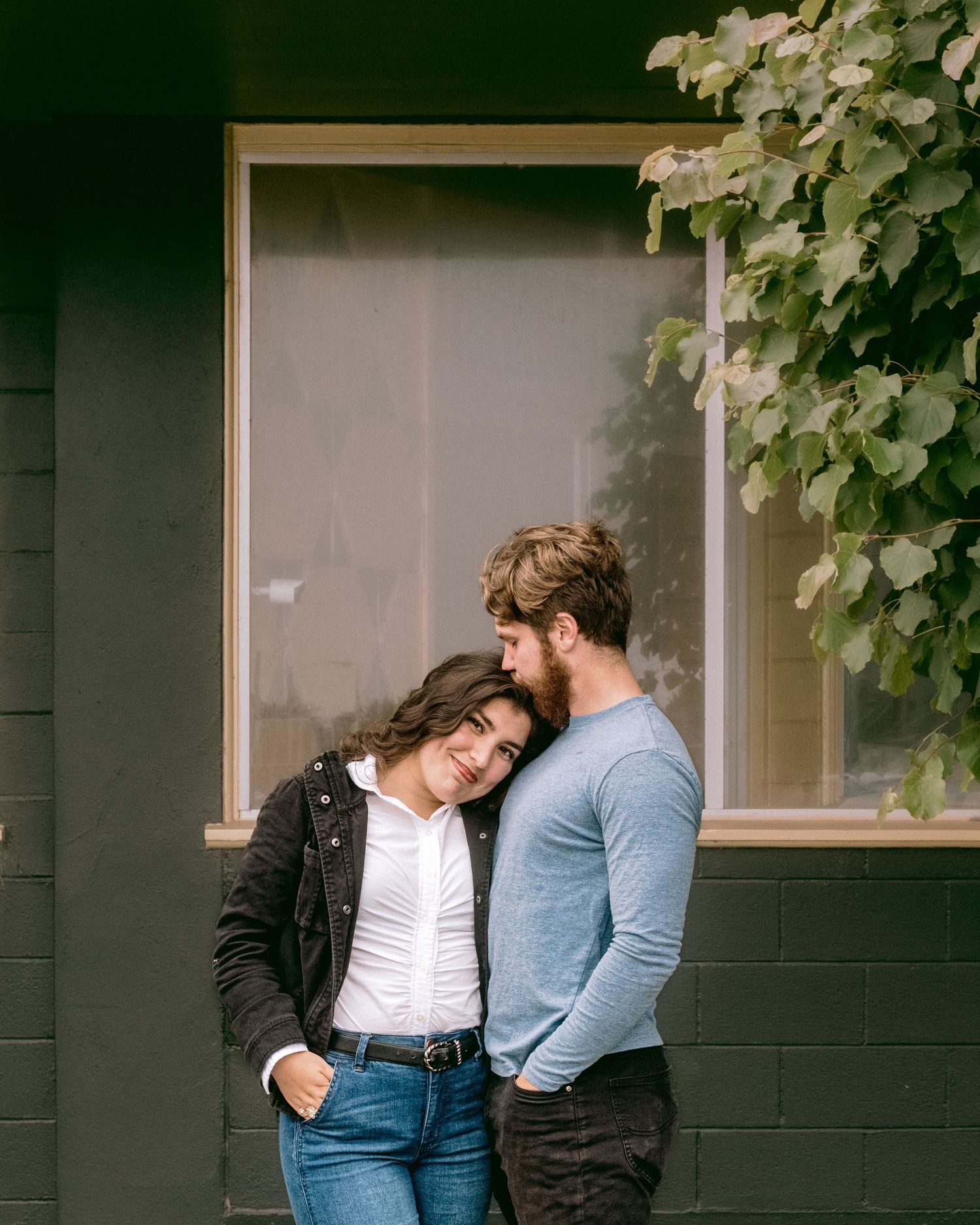 A couple shares an intimate moment against a dark wall with green foliage in the background.