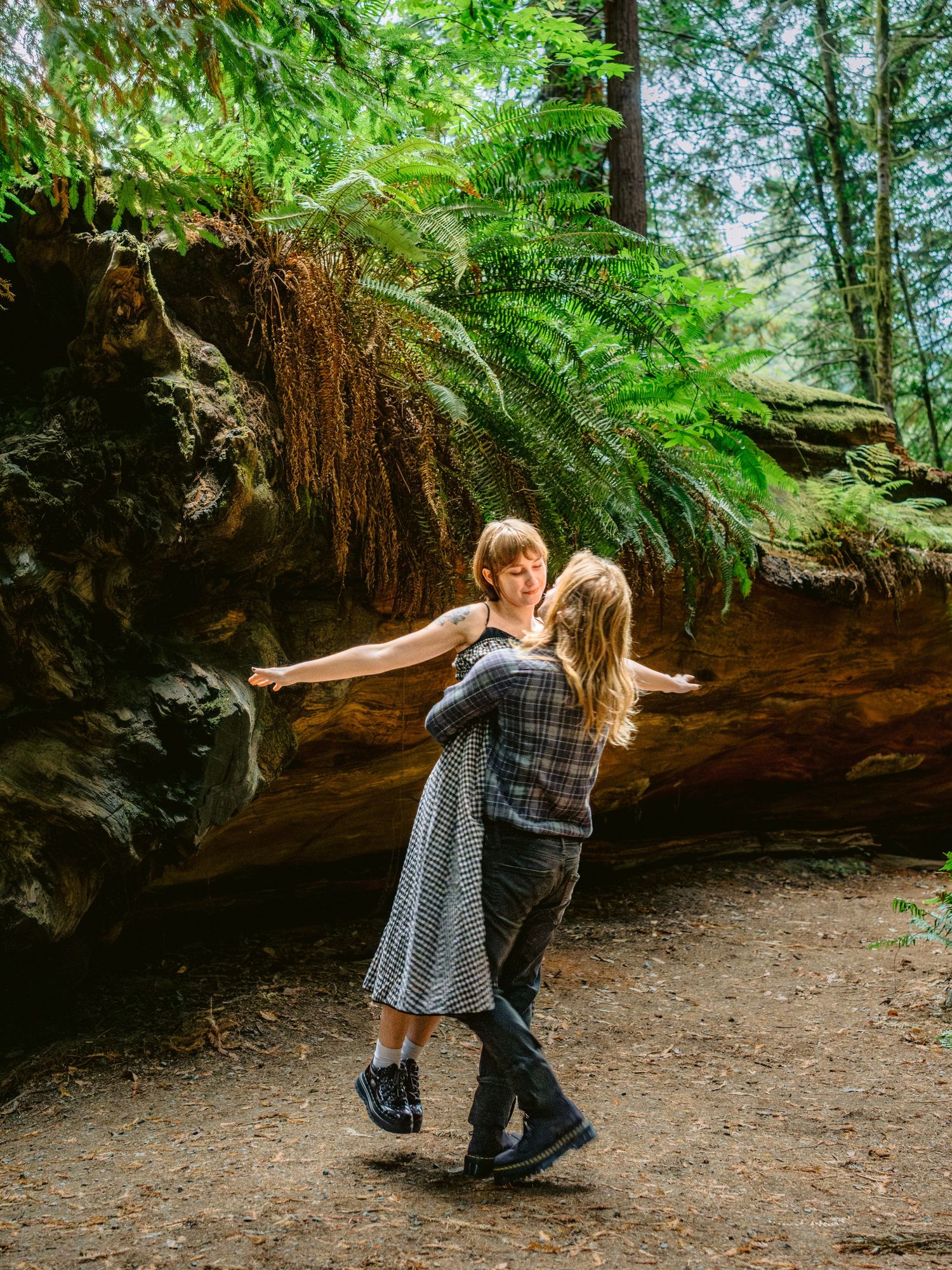 A couple dances playfully on a dirt path under tall trees and ferns.