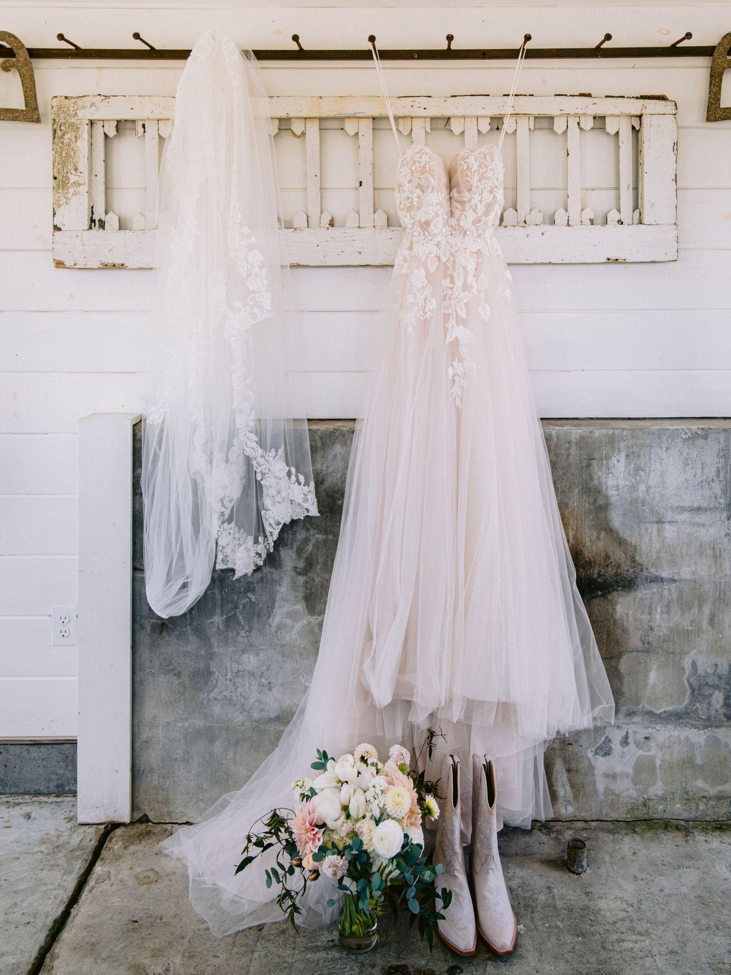 Wedding dress hanging against rustic white wall with floral bouquet below.