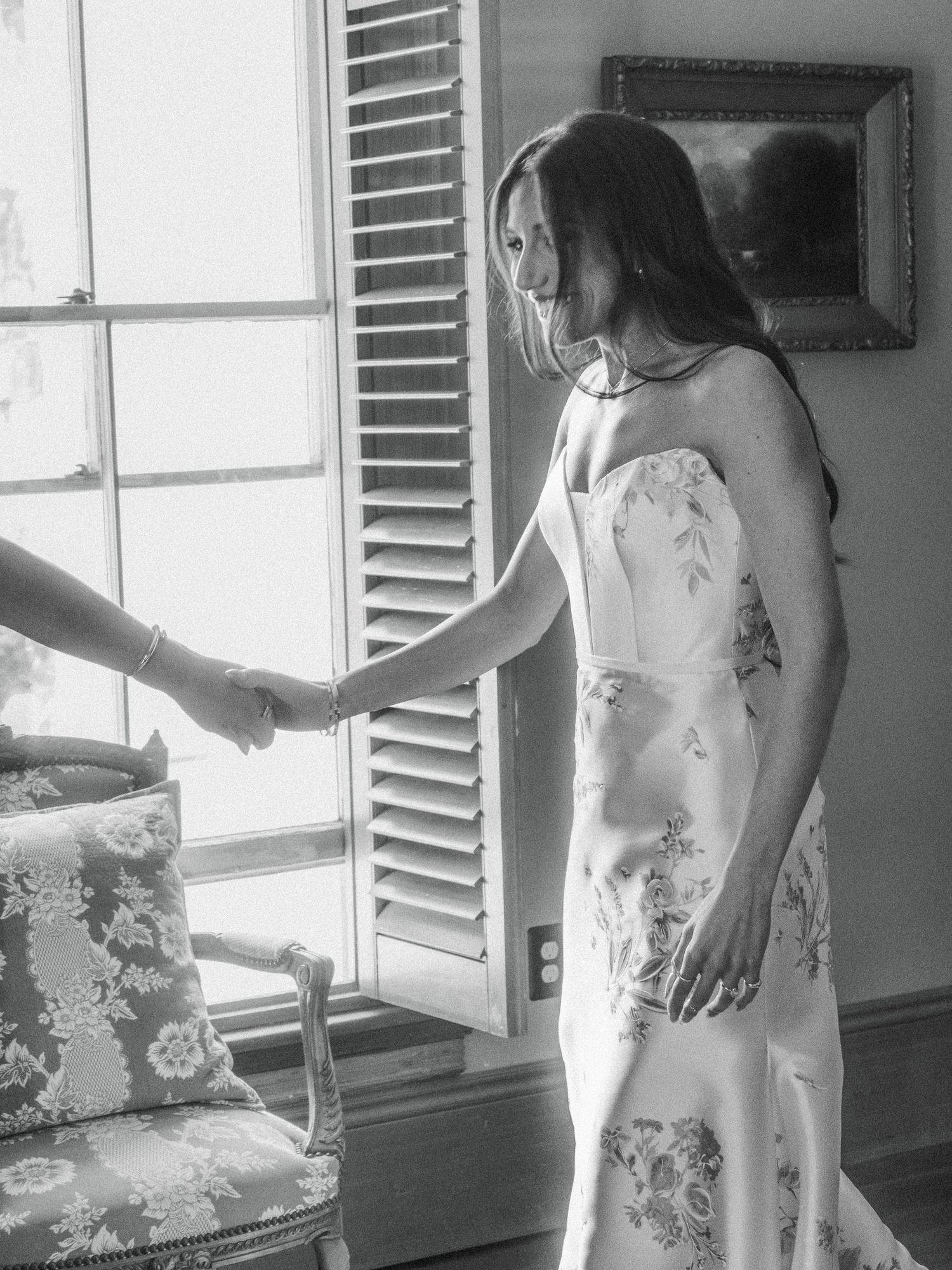 Black and white photo of a bride in a floral dress near a window with natural light.