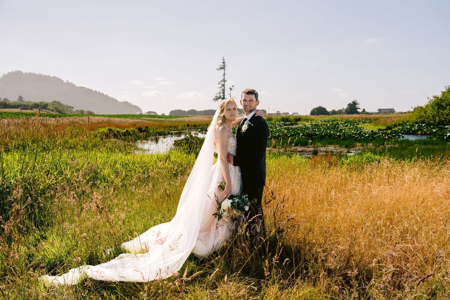 Wedding couple poses in a scenic field with mountains visible in the background.