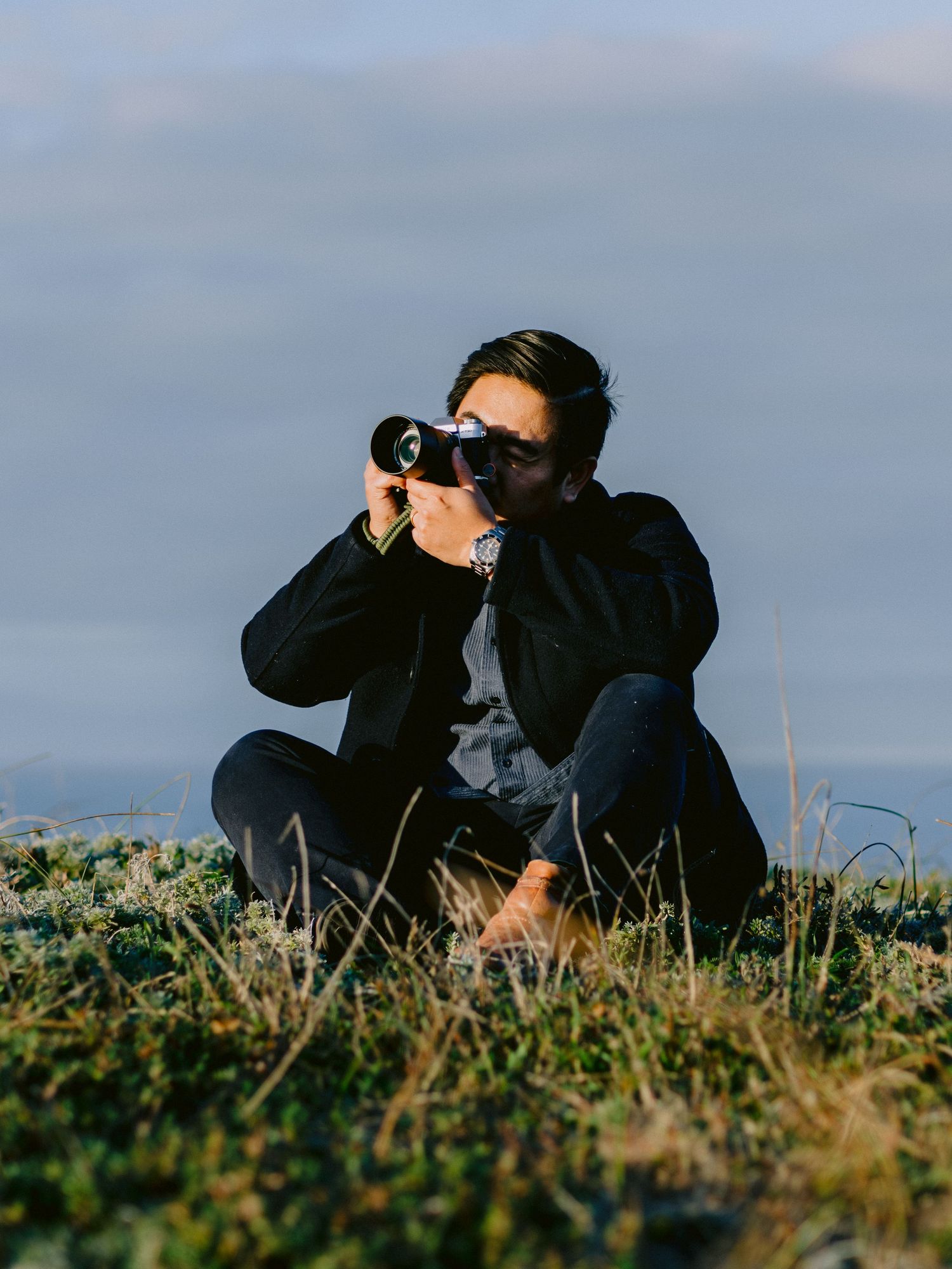 Person using binoculars to observe wildlife while sitting in grassy terrain against cloudy sky.