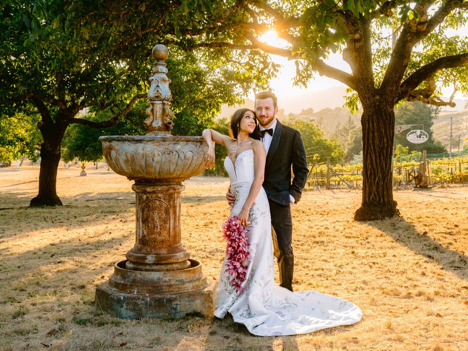 A wedding couple poses next to an ornate stone fountain in a garden during sunset with trees in the background.