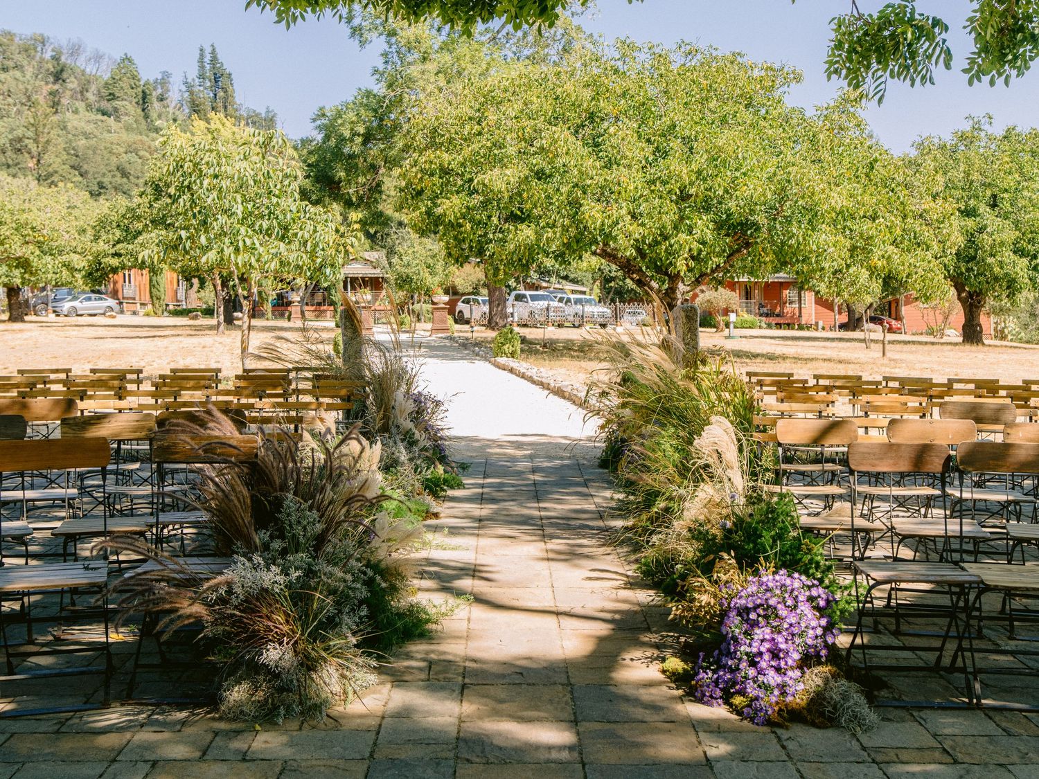 Rustic outdoor wedding venue with wooden benches arranged in rows under shady trees.
