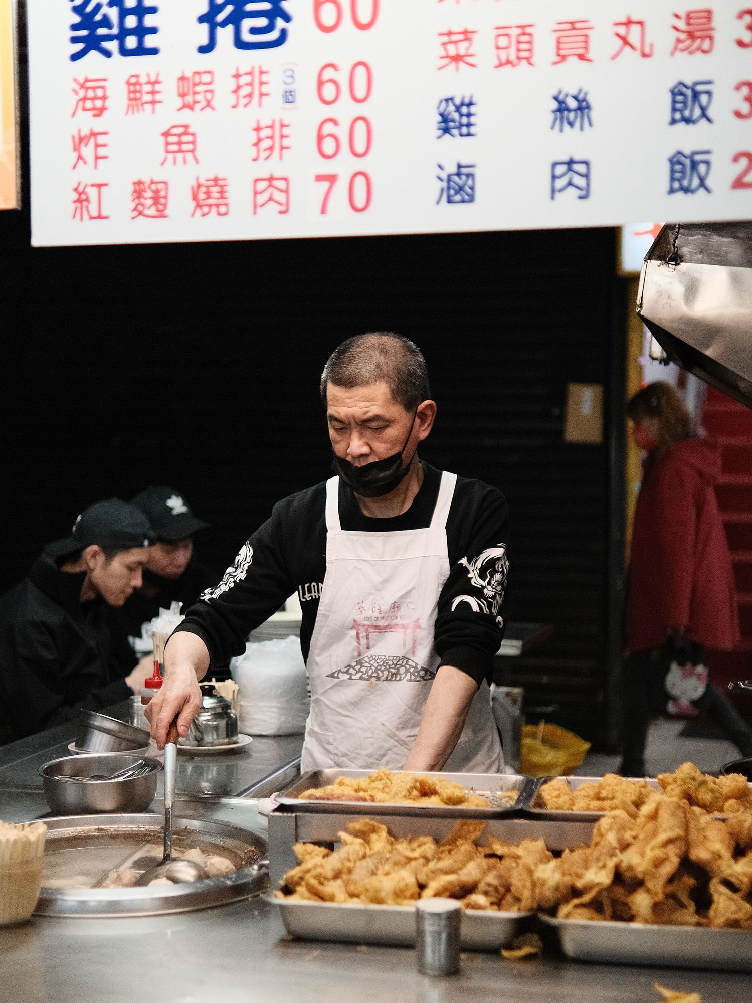 Chef preparing fried food in a commercial kitchen wearing black uniform and white apron.