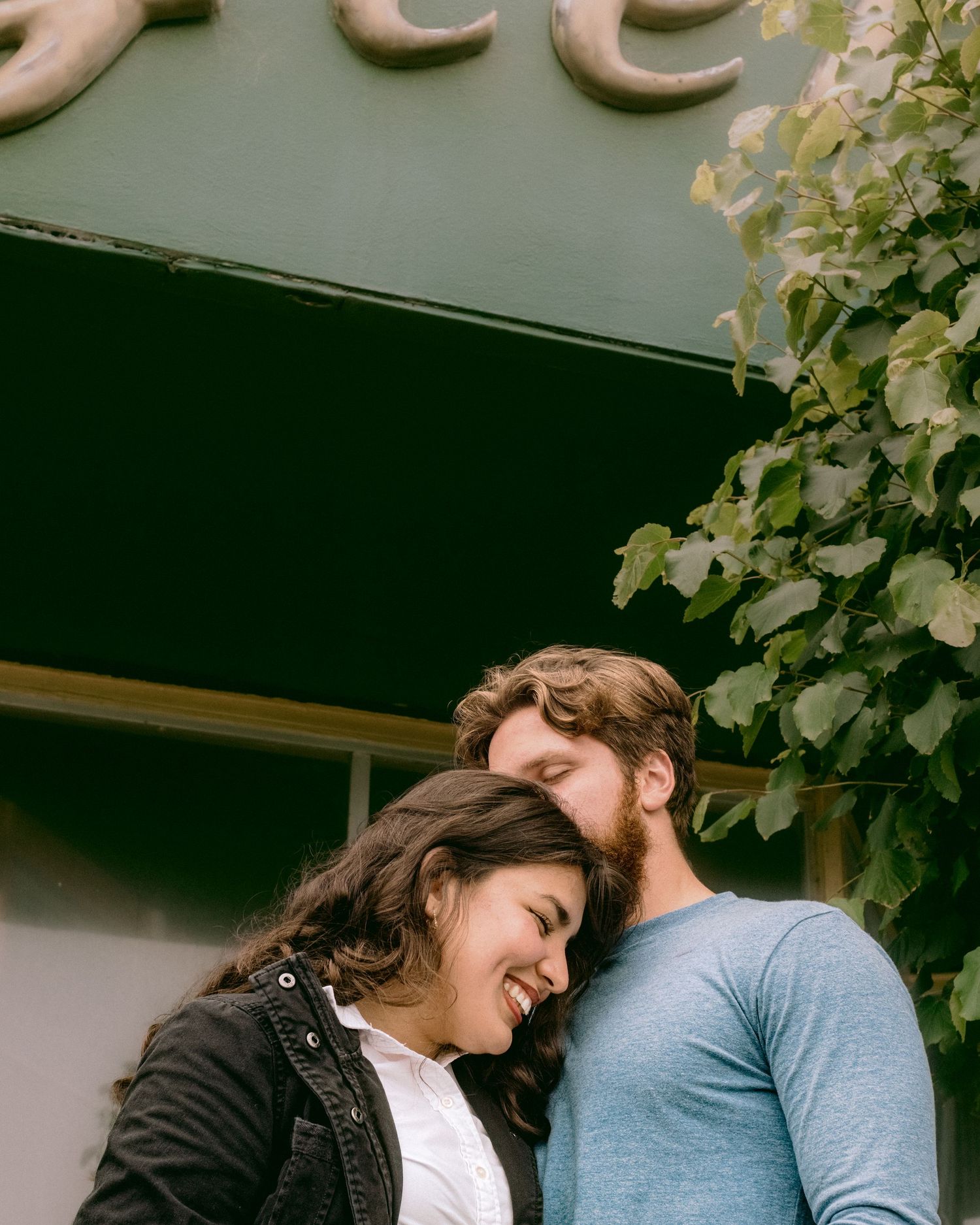A couple shares a tender moment while embracing outside a building with climbing vines.