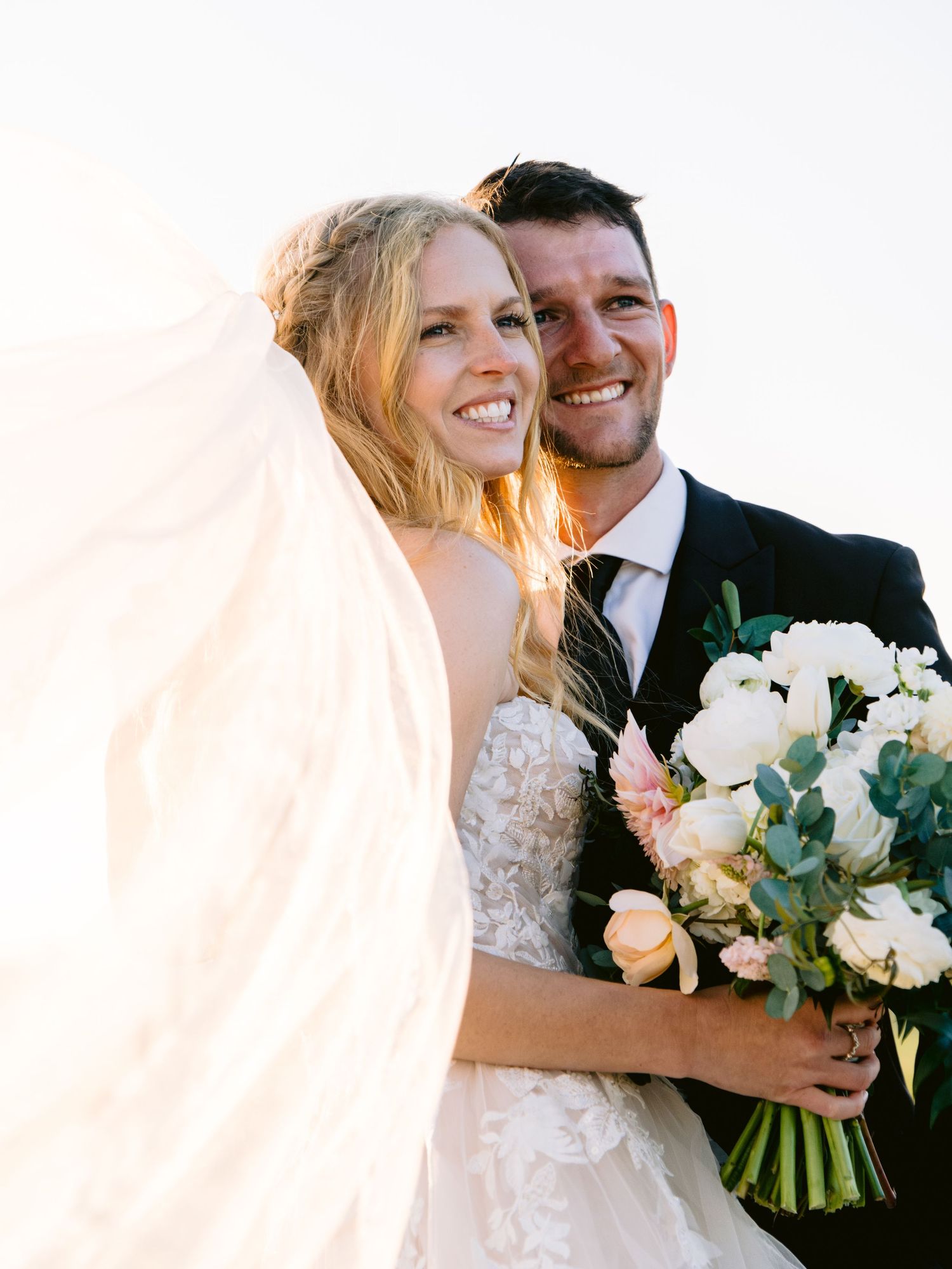 Newlyweds share intimate moments while holding a bouquet of white and peach flowers.