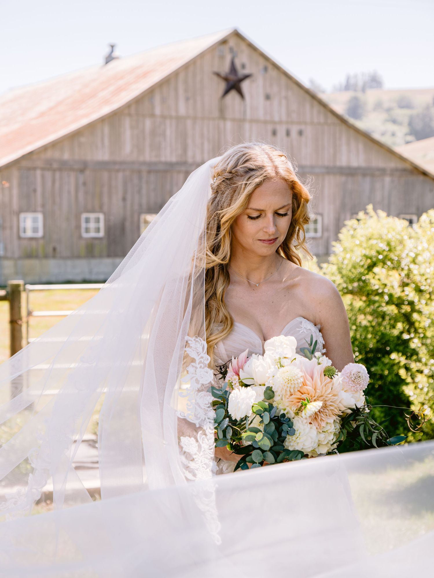 Bride with flowing veil poses near a rustic barn holding an elegant floral bouquet.