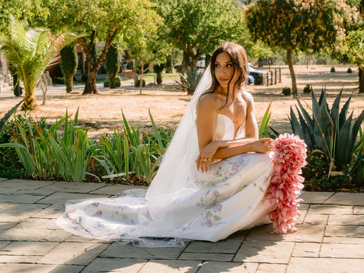 A bride in a flowing white dress sits elegantly with a pink bouquet in a garden setting.