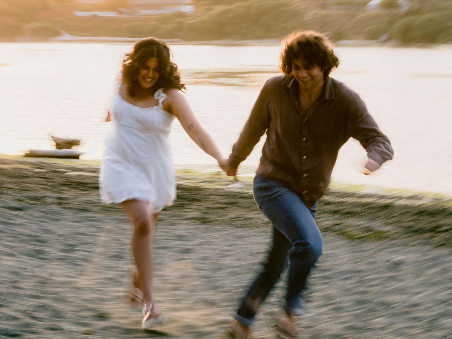 Two people run joyfully together on a beach at sunset holding hands.