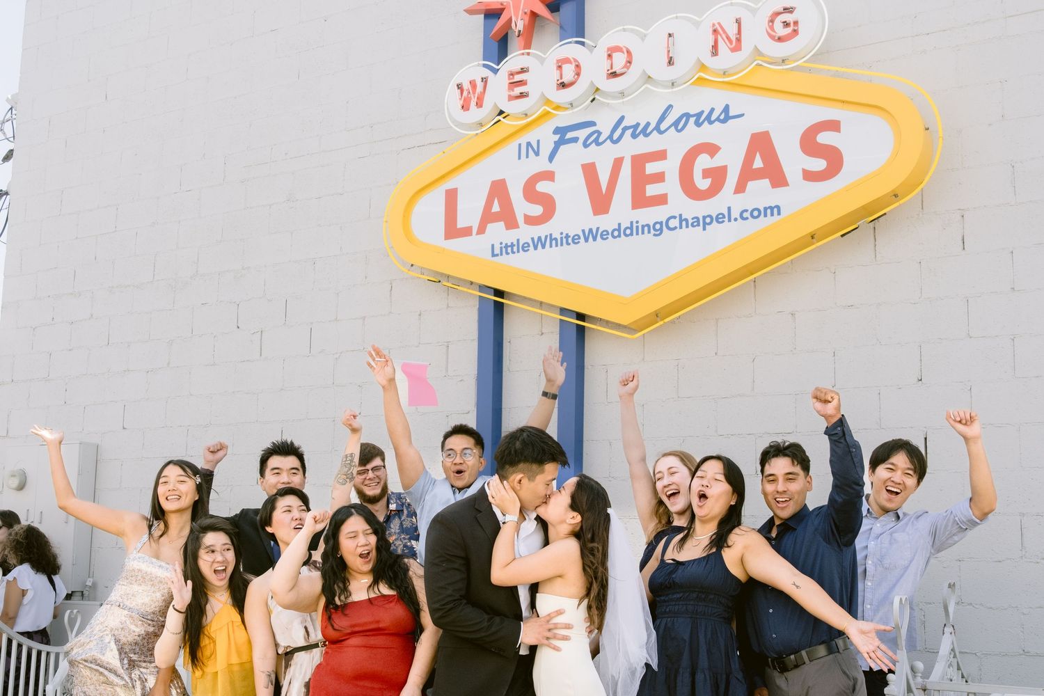 A large group celebrates together with raised arms in front of a Las Vegas wedding chapel sign.