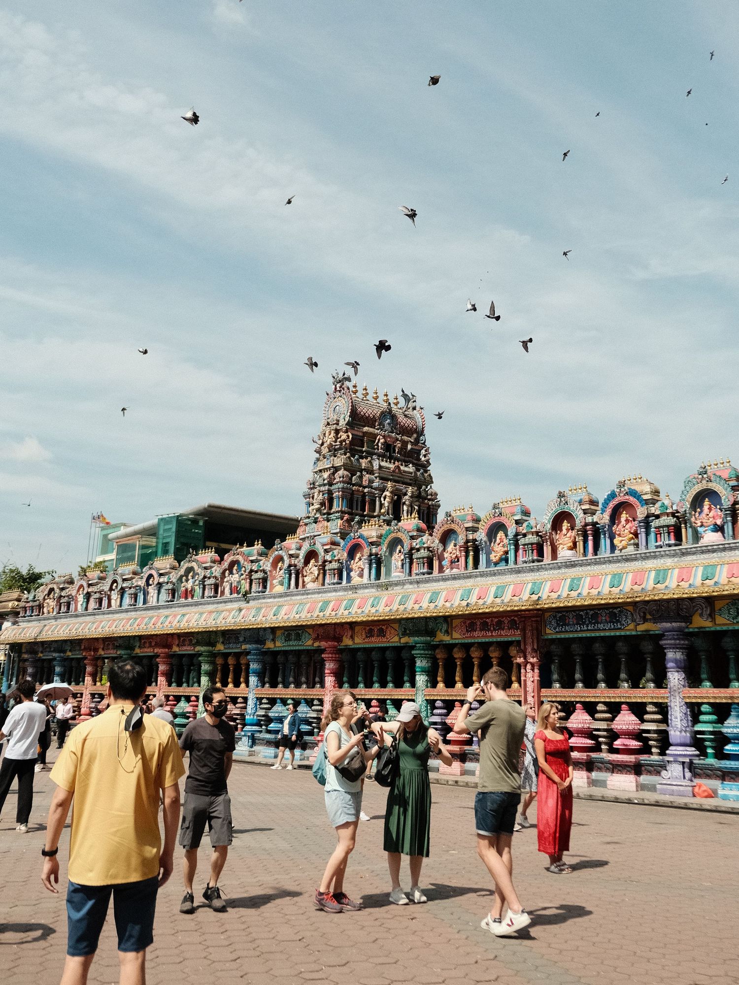 Busy plaza with ornate traditional Asian architecture and birds flying overhead.
