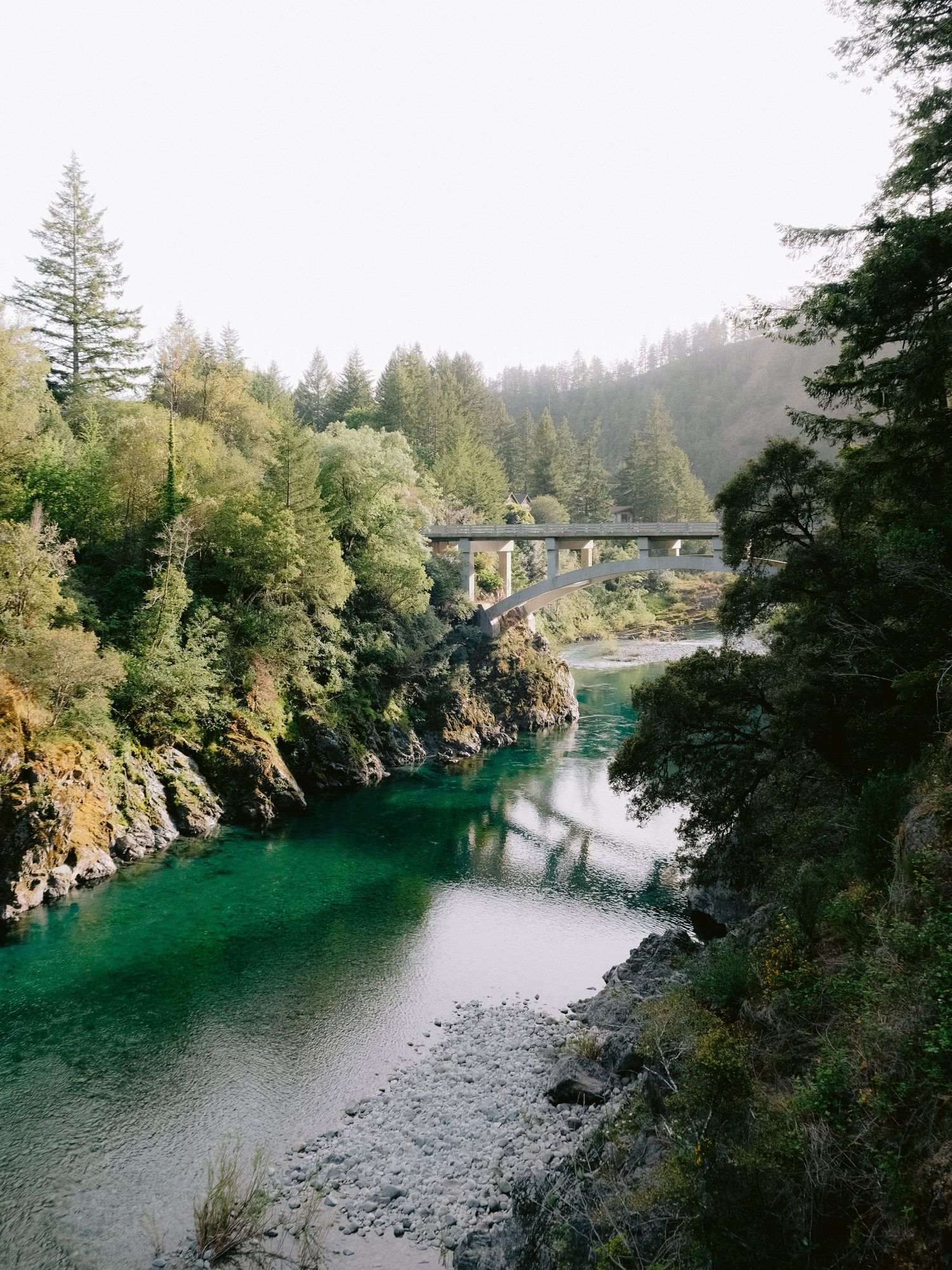 Scenic river view with emerald green water flowing under a bridge surrounded by forest.