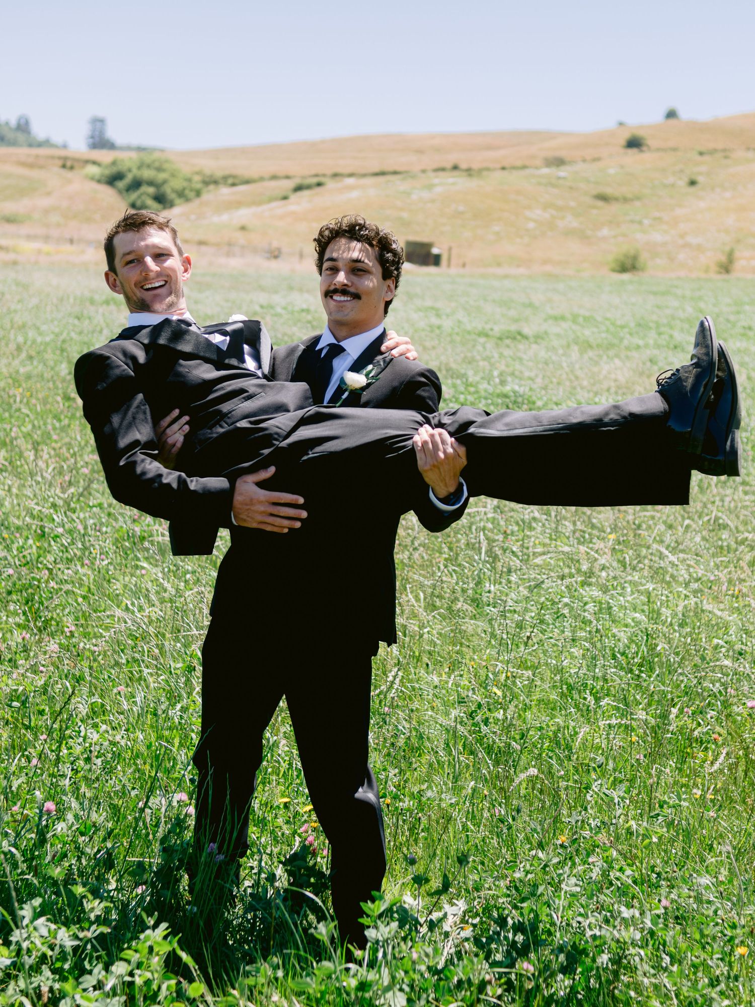 Groomsmen in black suits perform a fun balancing pose in a grassy field on a sunny day.