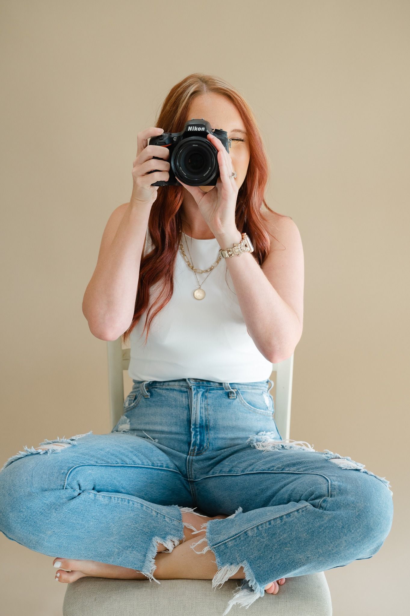 Person in white top and distressed jeans sitting cross-legged while holding a camera for self portraits.