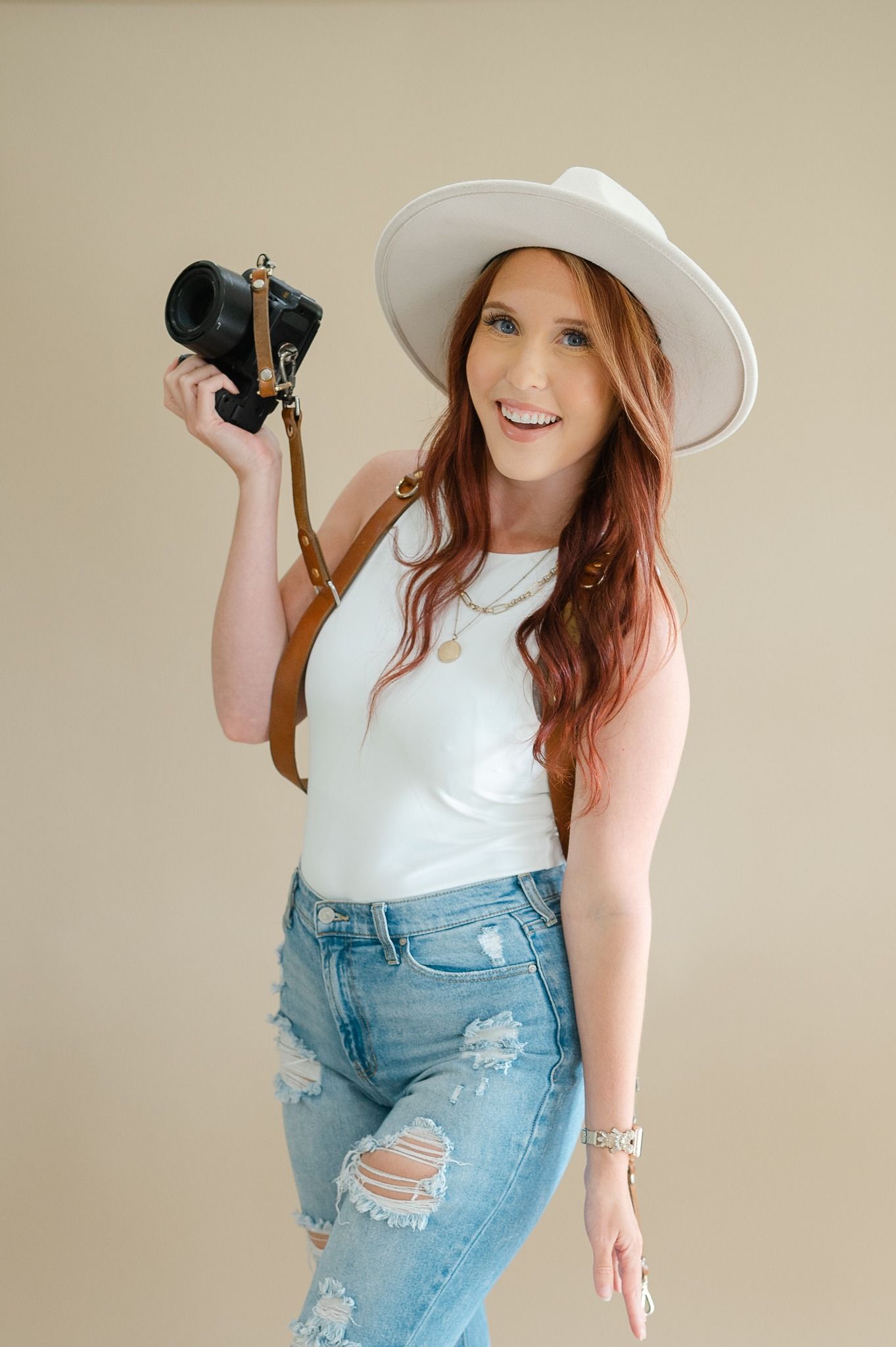 Woman in white sunhat and sleeveless top poses playfully with distressed denim jeans against neutral background.